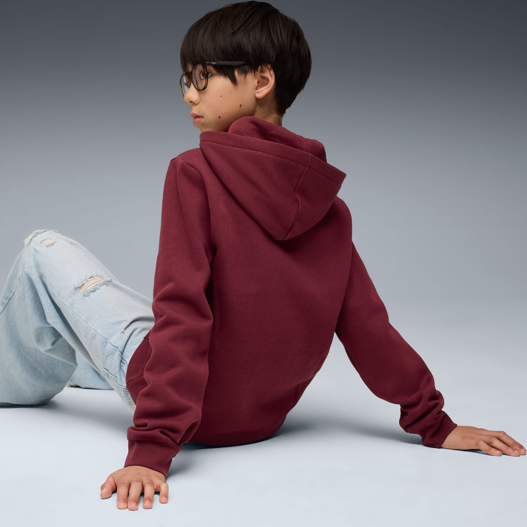 Een model zit tegen een grijze achtergrond, gekleed in een bordeauxrode hoodie en gescheurde jeans.
