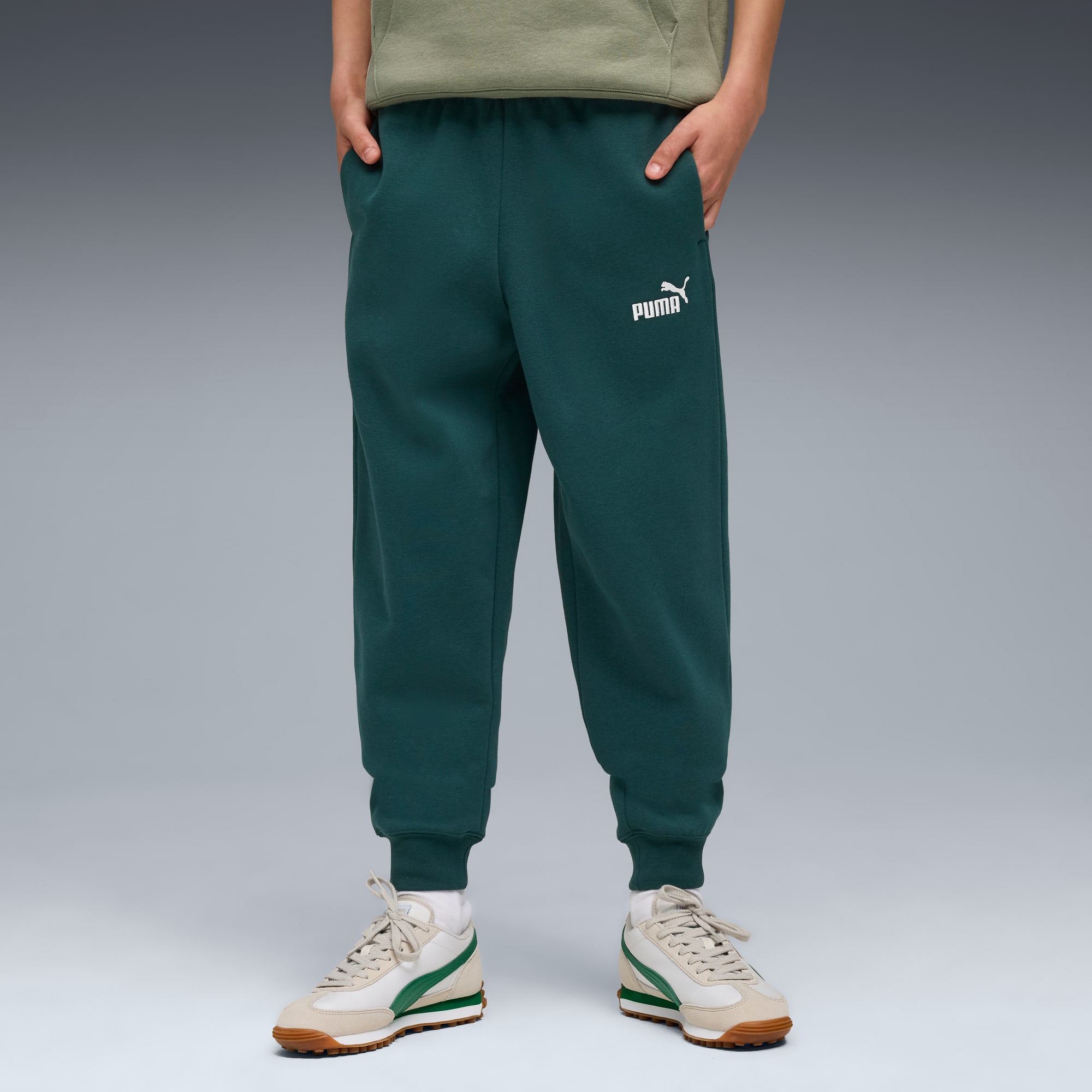 Ein Model posiert vor grauem Hintergrund mit grüner Jogginghose mit Puma-Logo und Turnschuhen.