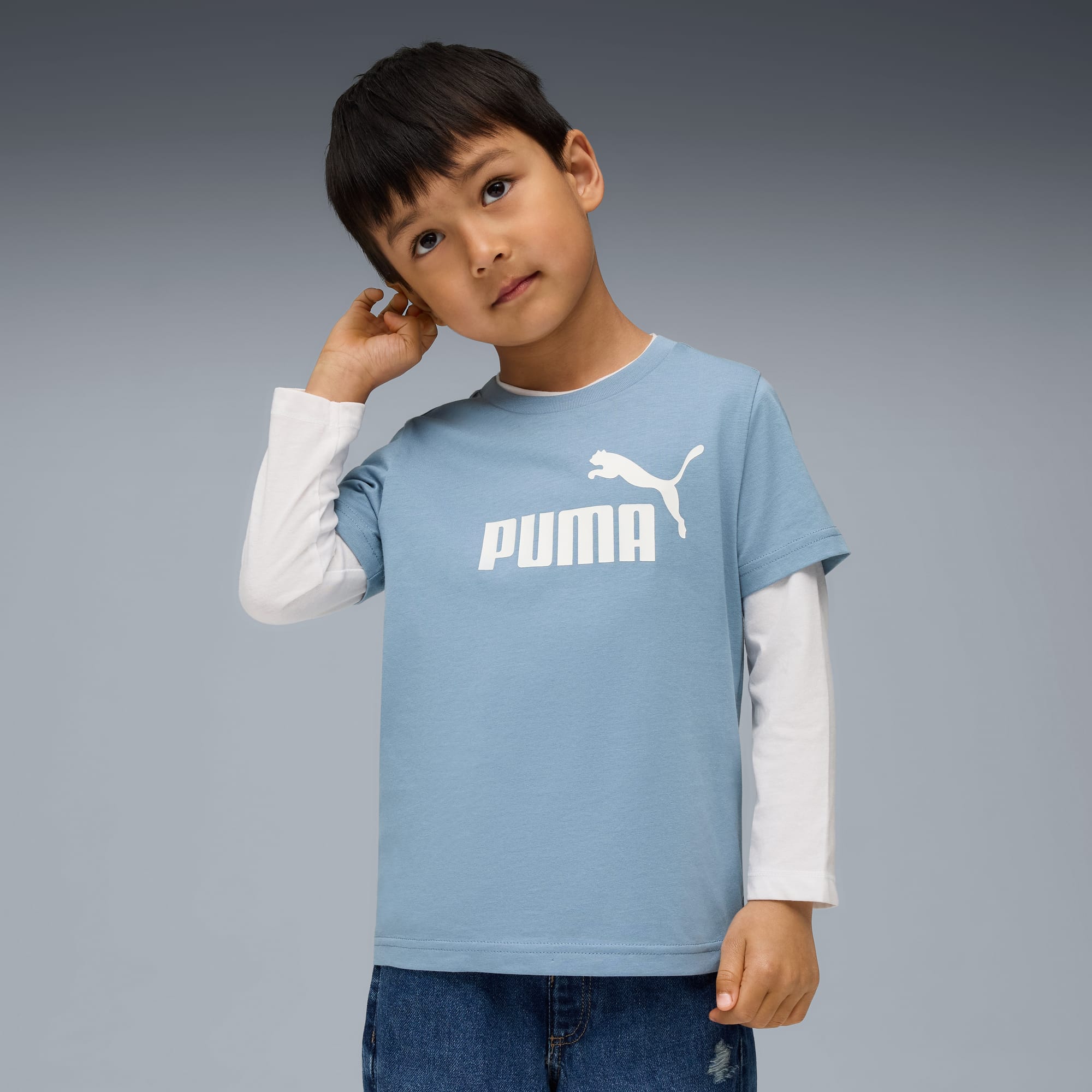 Ein junger Junge posiert vor grauem Hintergrund und trägt ein blaues Puma-T-Shirt und Jeans.