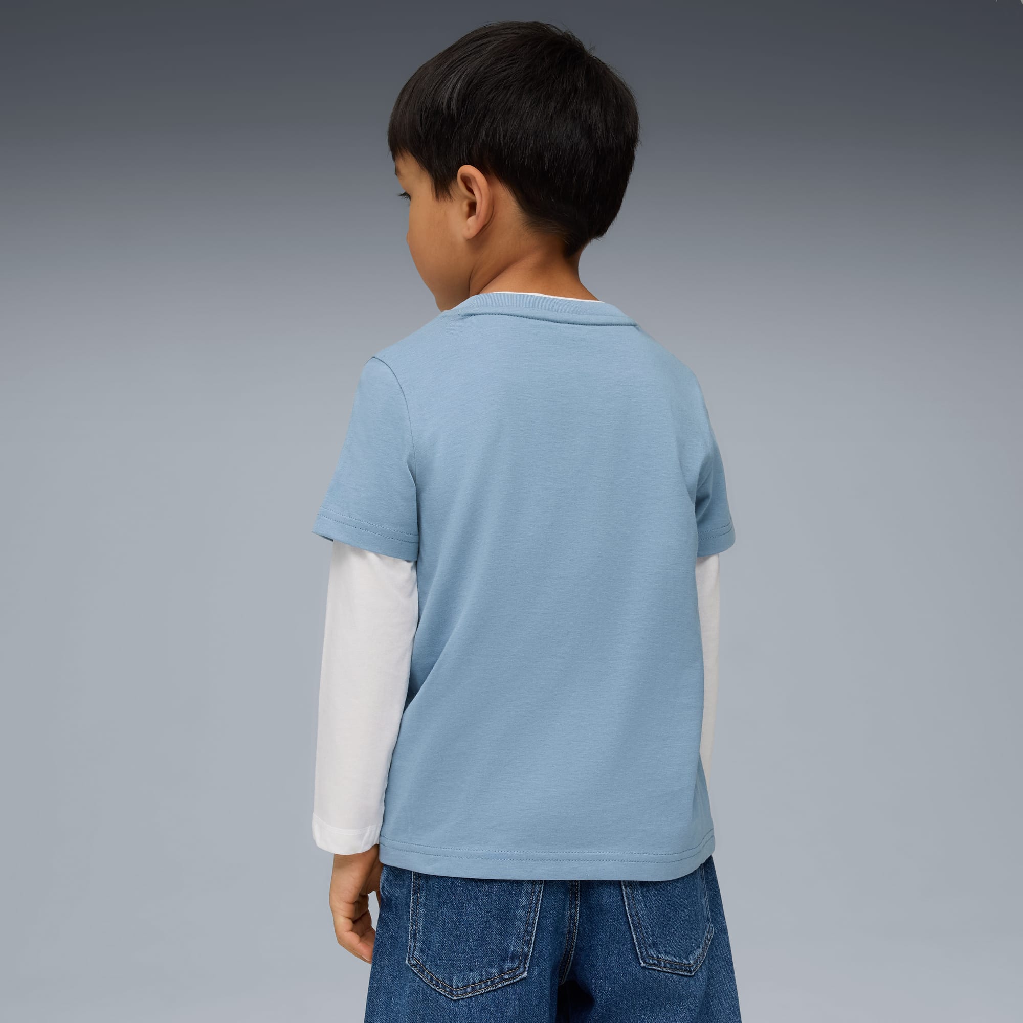 Ein Model mit dunklen Haaren, blauem T-Shirt, weißen Ärmeln und Jeans steht vor grauem Hintergrund.