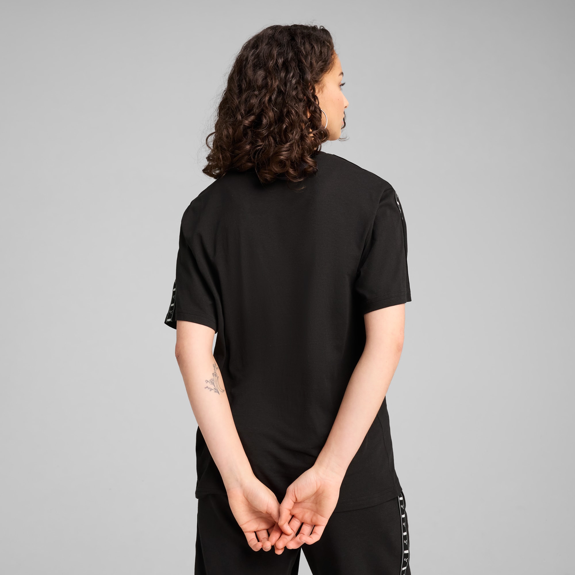 Model met krullend haar staat met de rug naar de camera, draagt zwart T-shirt, grijze achtergrond.