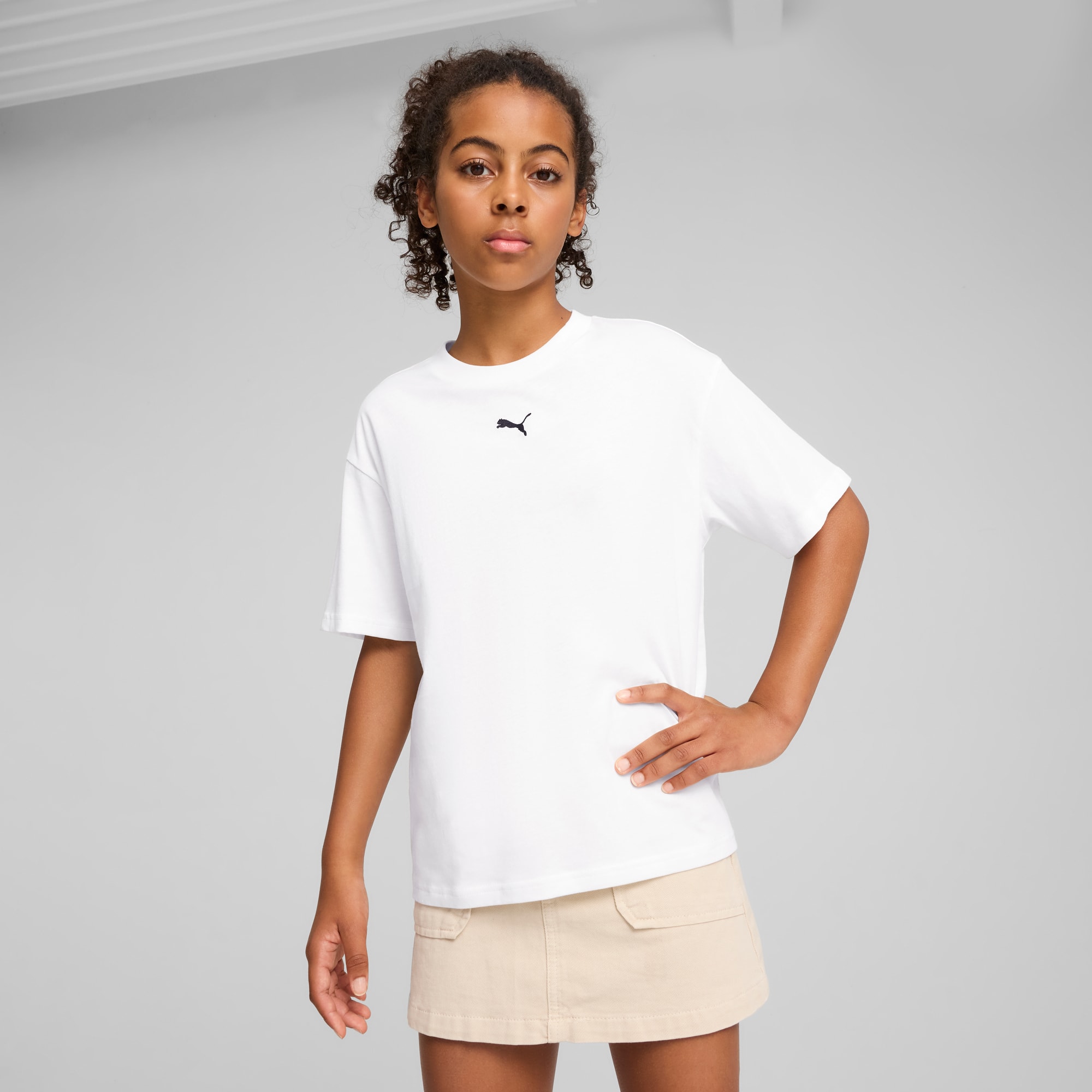 Een model poseert tegen een grijze achtergrond, gekleed in een wit T-shirt en beige rok.