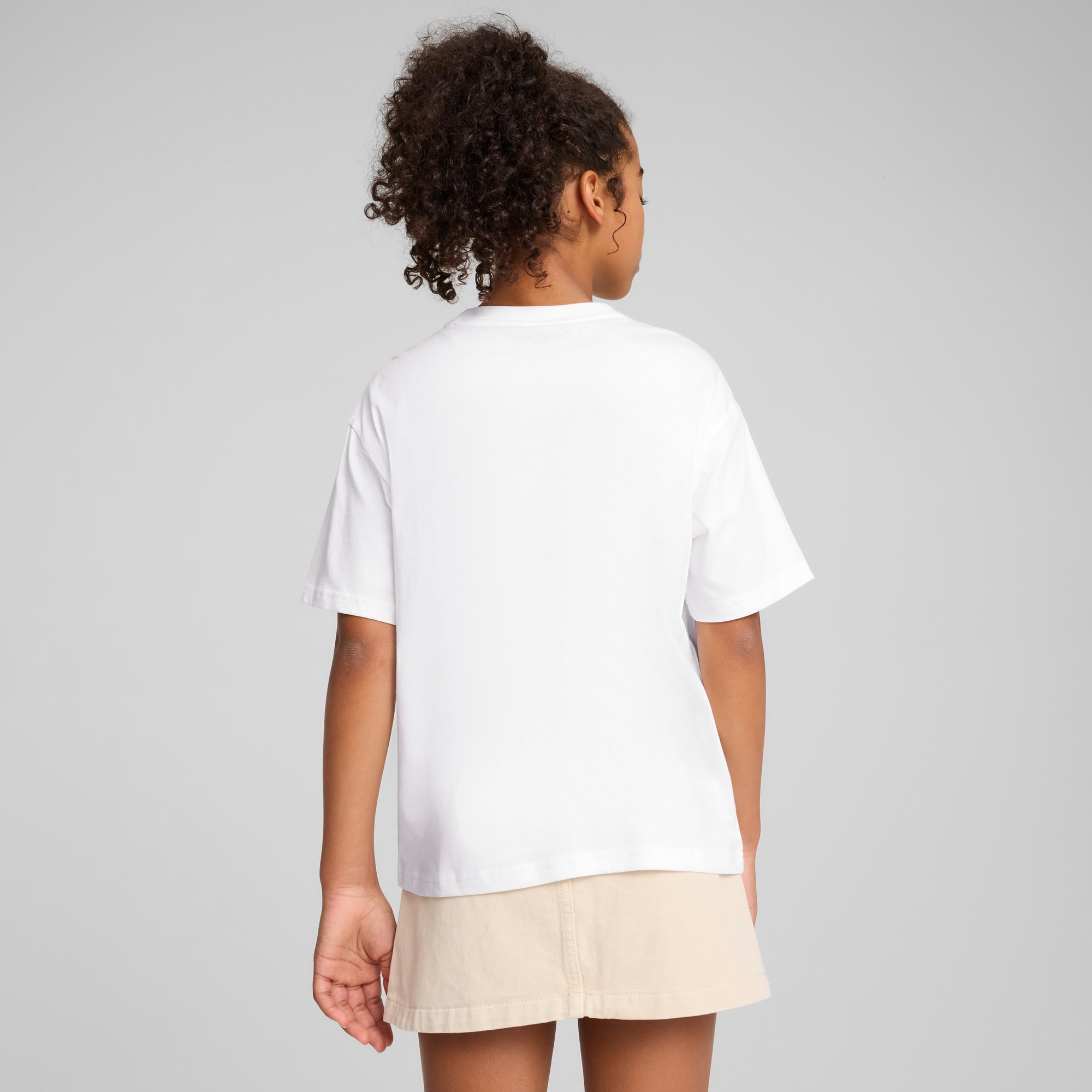 Ein Model steht vor grauem Hintergrund, trägt ein weißes T-Shirt und hellbraunen Rock, Rückansicht.