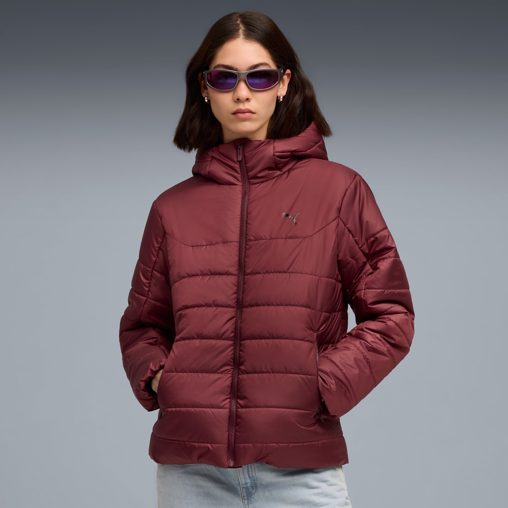 Een model poseert tegen een grijze achtergrond met een kastanjebruine pufferjas en zonnebril.
