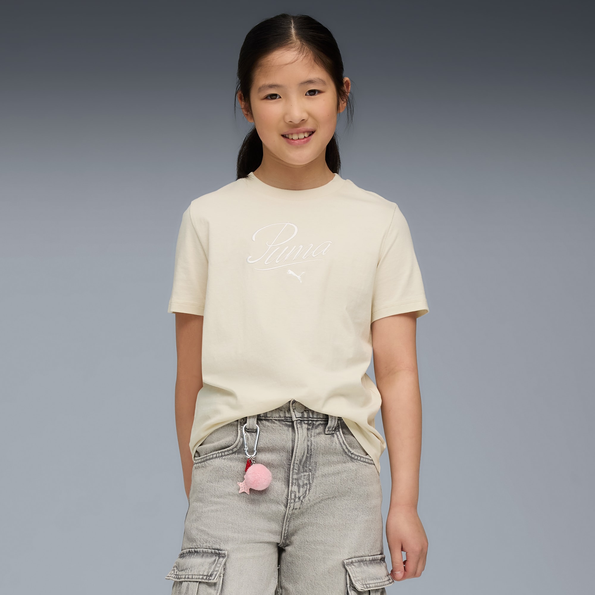 Una bambina sorridente posa su sfondo grigio con una maglietta color crema, pantaloncini grigi e un pon pon.