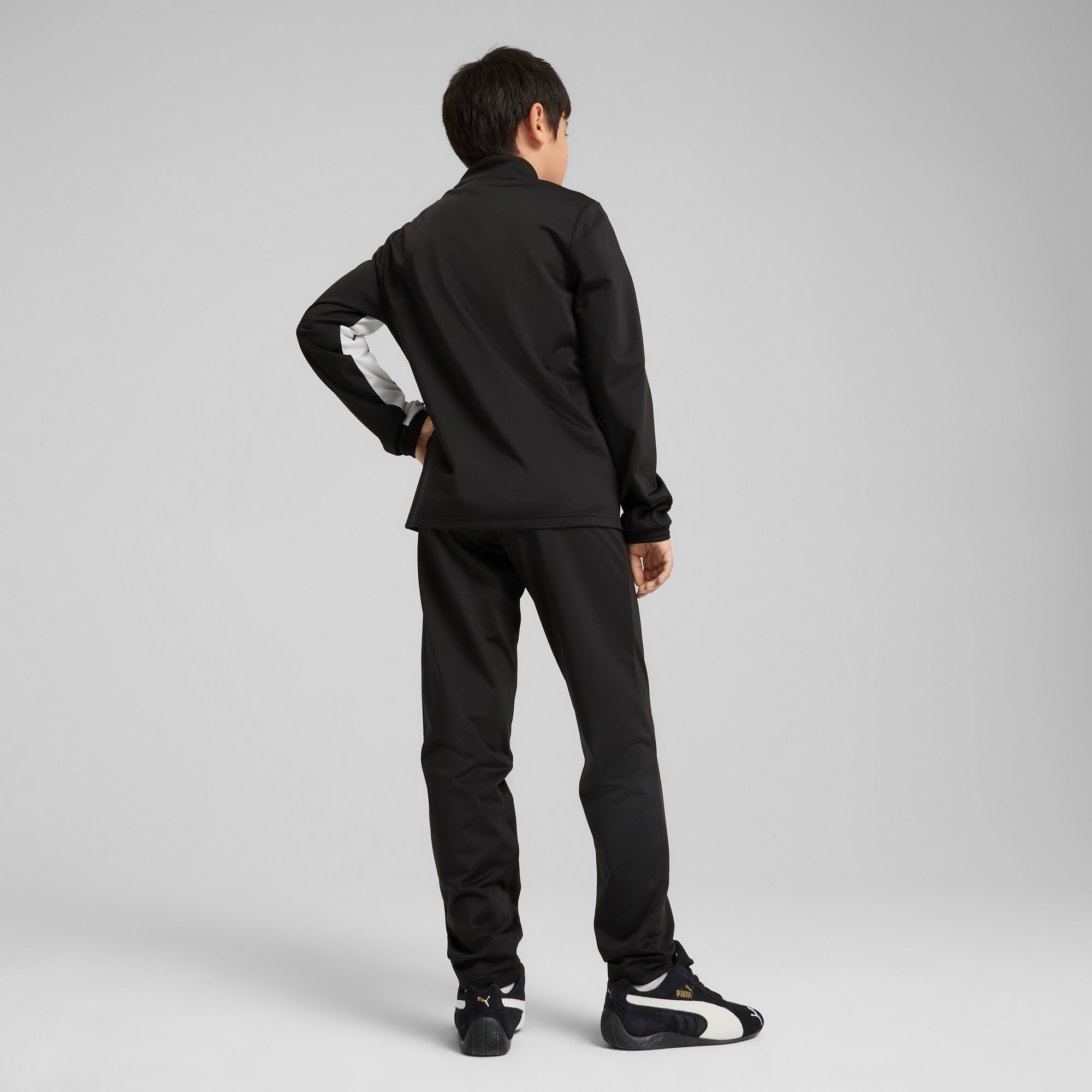 Ein Model steht vor grauem Hintergrund und trägt einen schwarzen Trainingsanzug mit weißen Details und Turnschuhen.