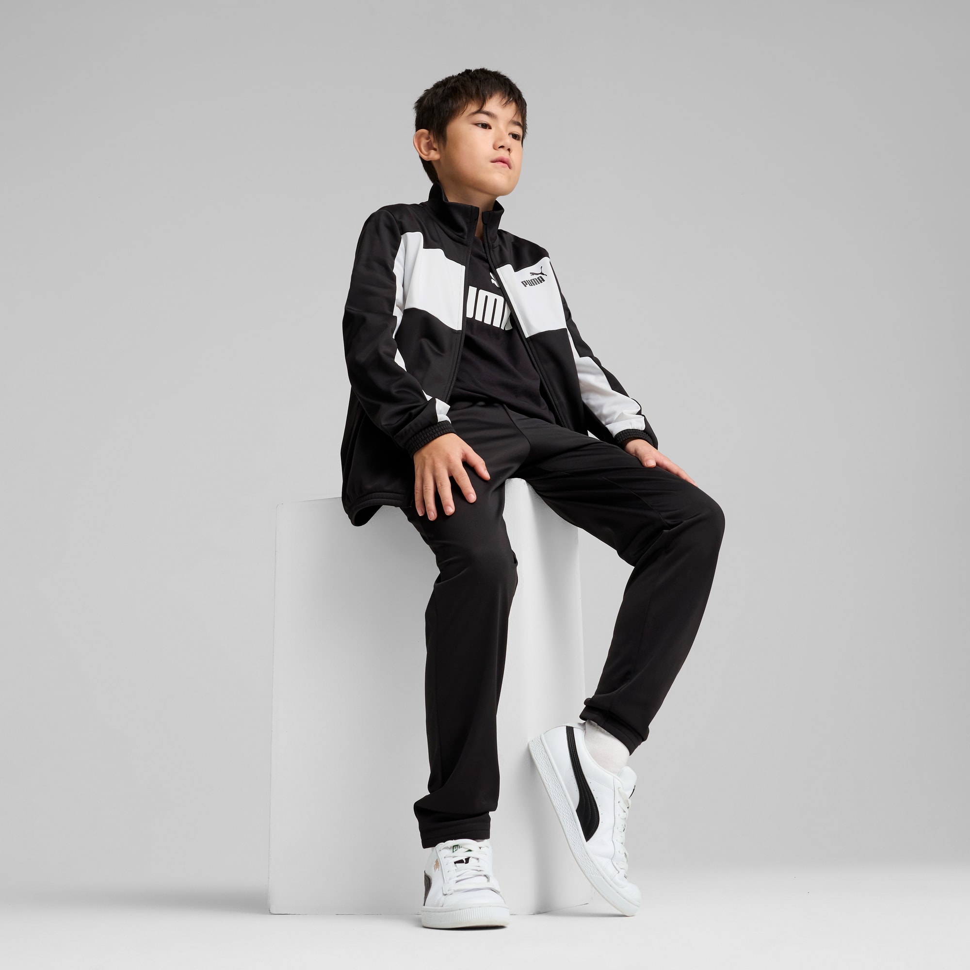 Na szarym tle model siedzi w dresie Puma oraz białych i czarnych butach sportowych.