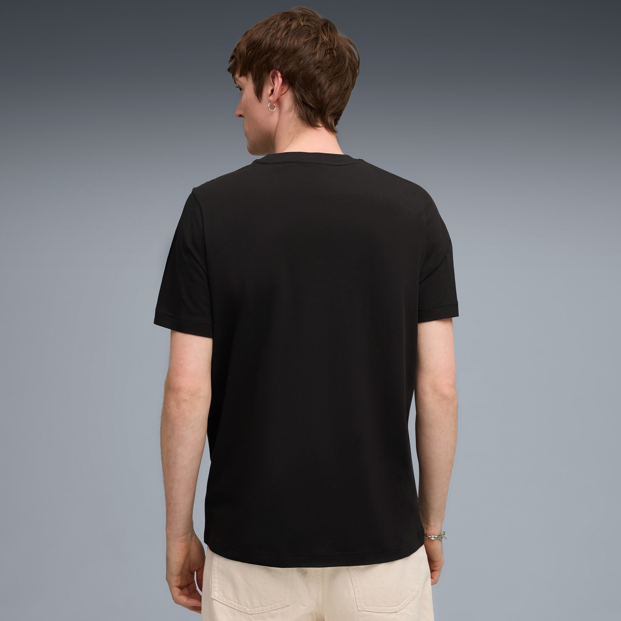 Een model staat met de rug naar de camera op grijs, met zwart shirt en lichte broek.