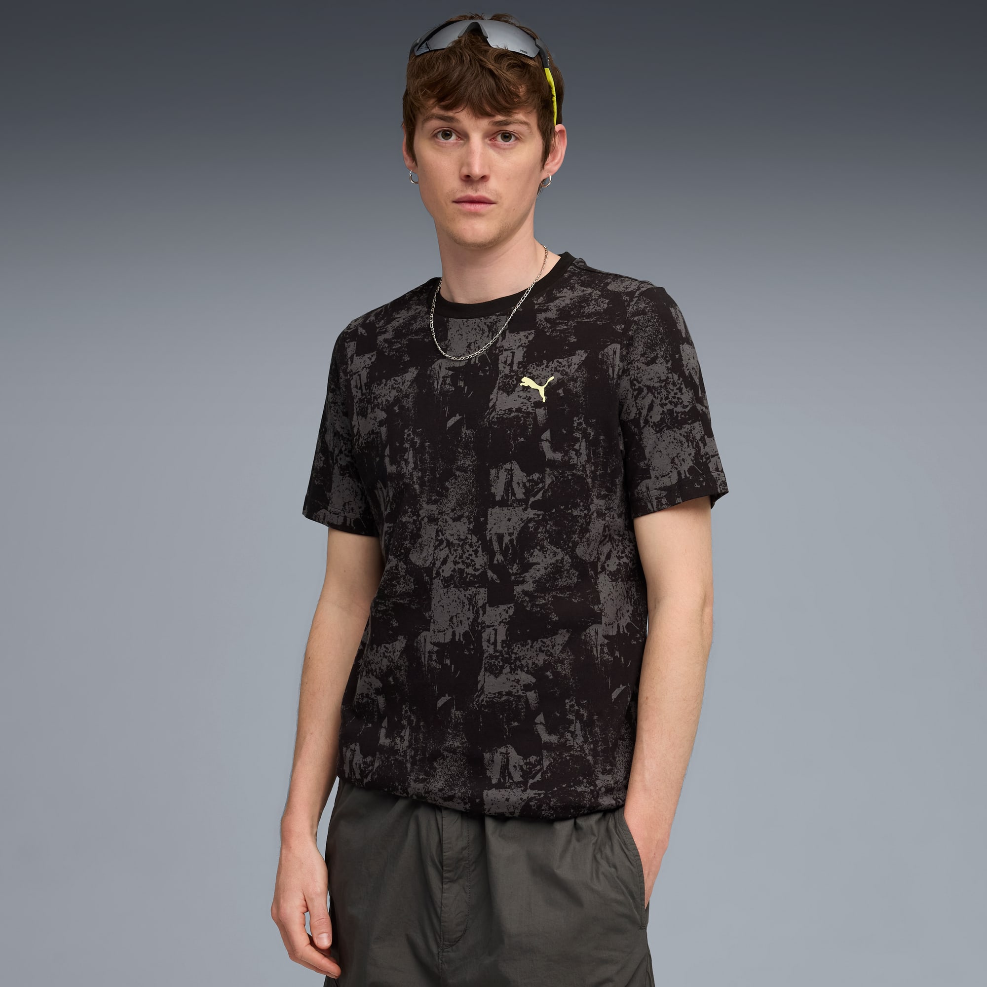 Ein Model posiert vor grauem Hintergrund, mit gemustertem T-Shirt und dunklen Shorts.