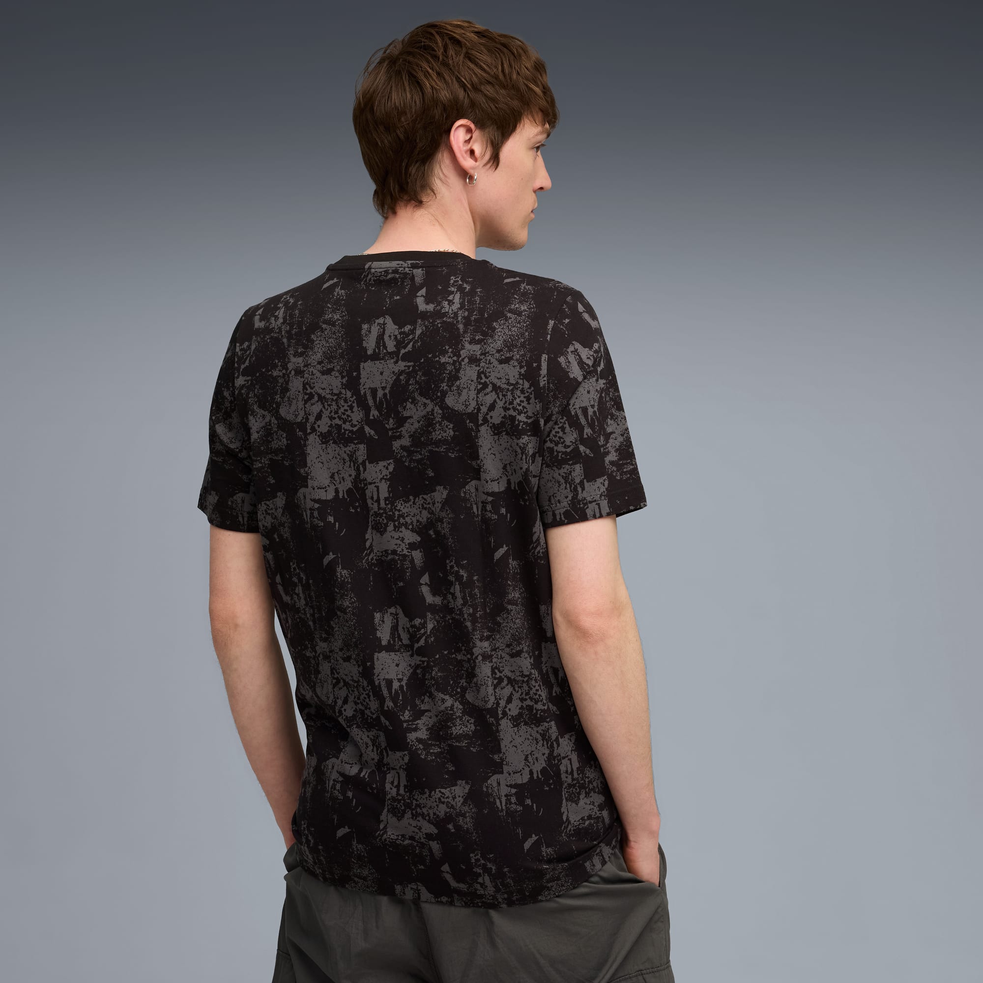 Ein Model posiert vor grauem Hintergrund mit gemustertem schwarzem T-Shirt und grauen Shorts.