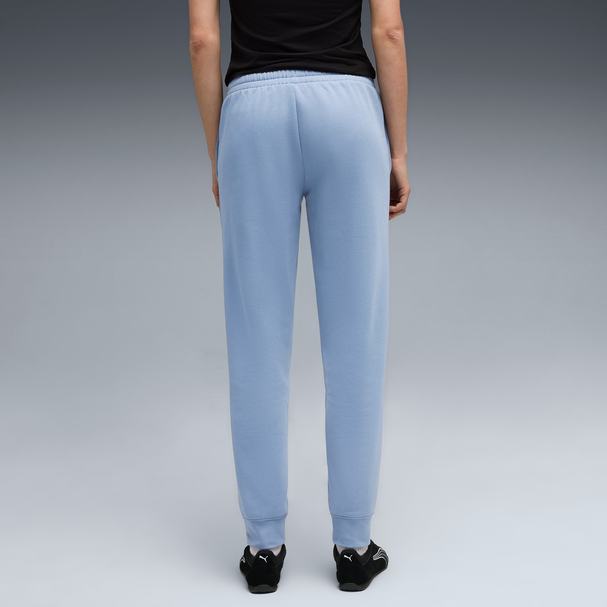 Een model staat op een grijze achtergrond, gekleed in een zwarte top, lichtblauwe broek en zwarte schoenen.