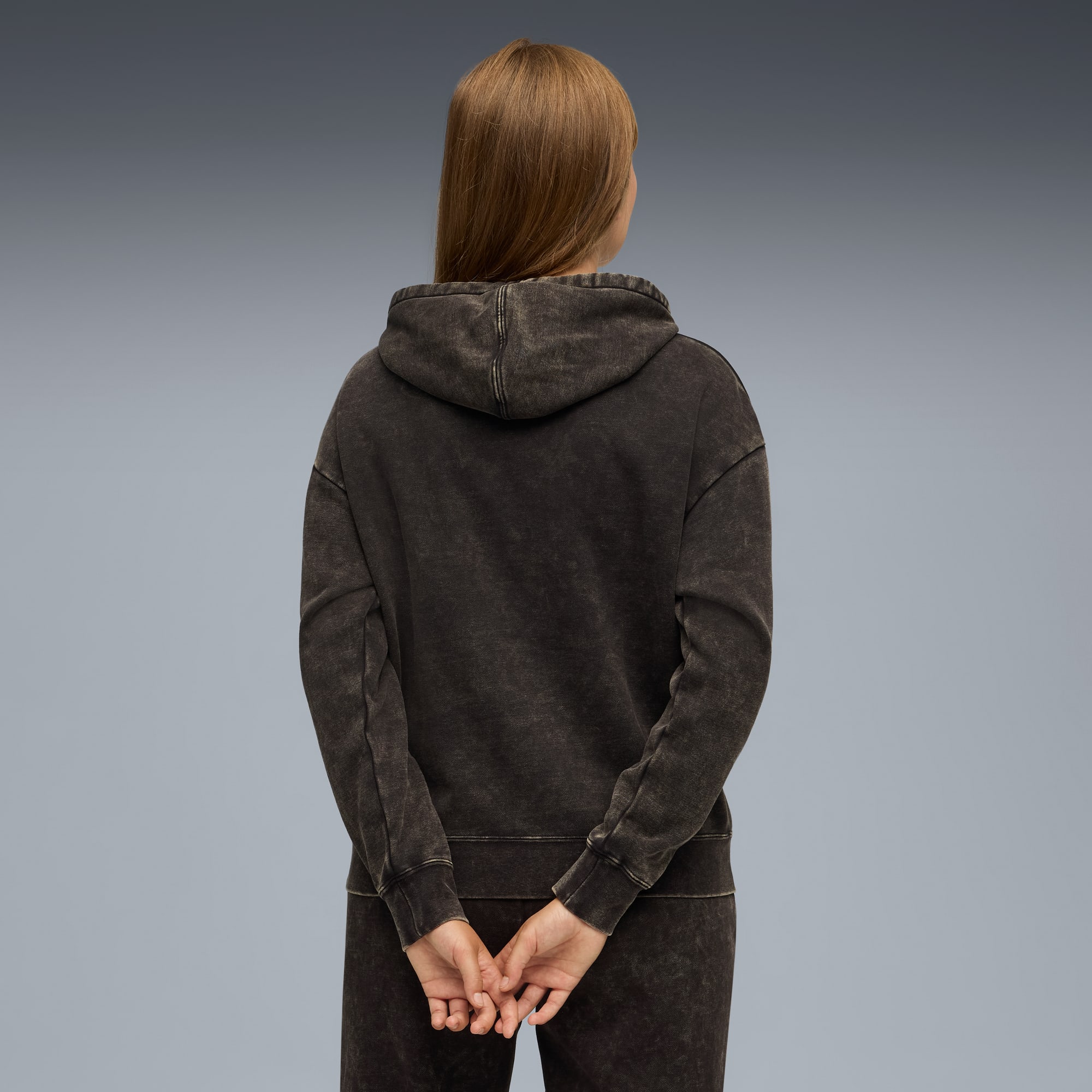 Vue de dos, une personne porte un sweat à capuche et un pantalon foncés sur fond gris.