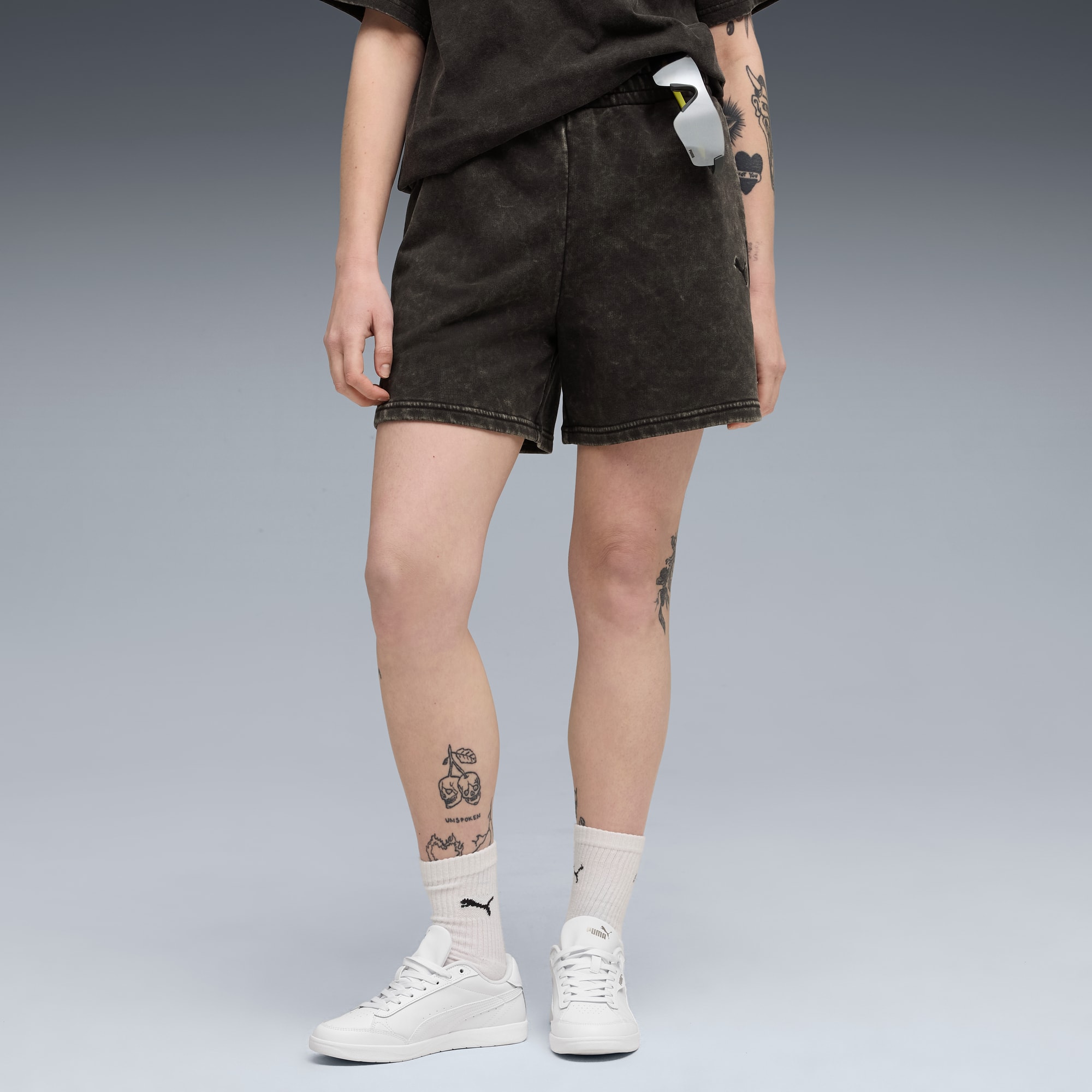 Sur fond gris, un modèle porte short foncé, haut, chaussettes blanches et baskets, montrant des tatouages sur les jambes.