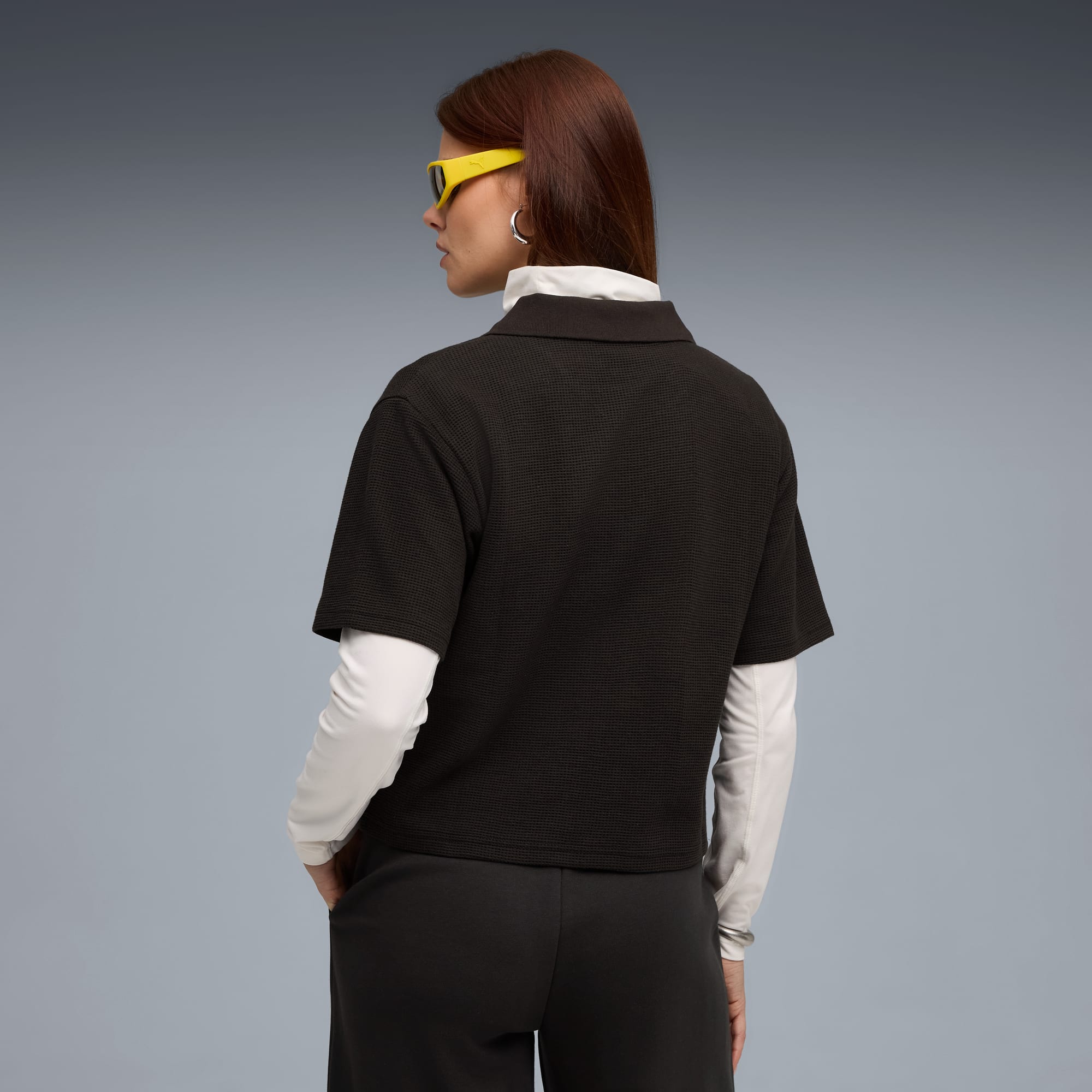 Su sfondo grigio, una modella di spalle indossa polo scura, top chiaro, pantaloni scuri e occhiali gialli.