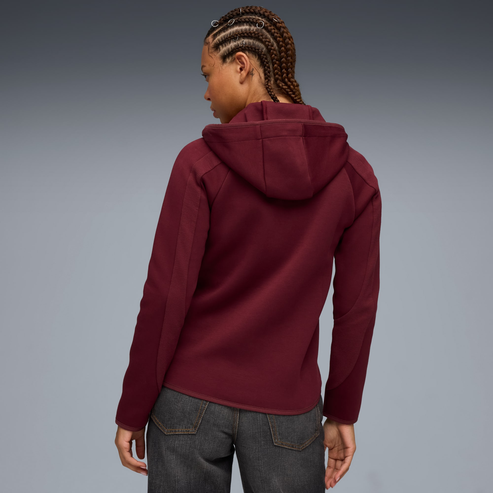 Een model staat met de rug naar de camera, tegen een grijze achtergrond, in een bordeauxrode hoodie en donkere jeans.