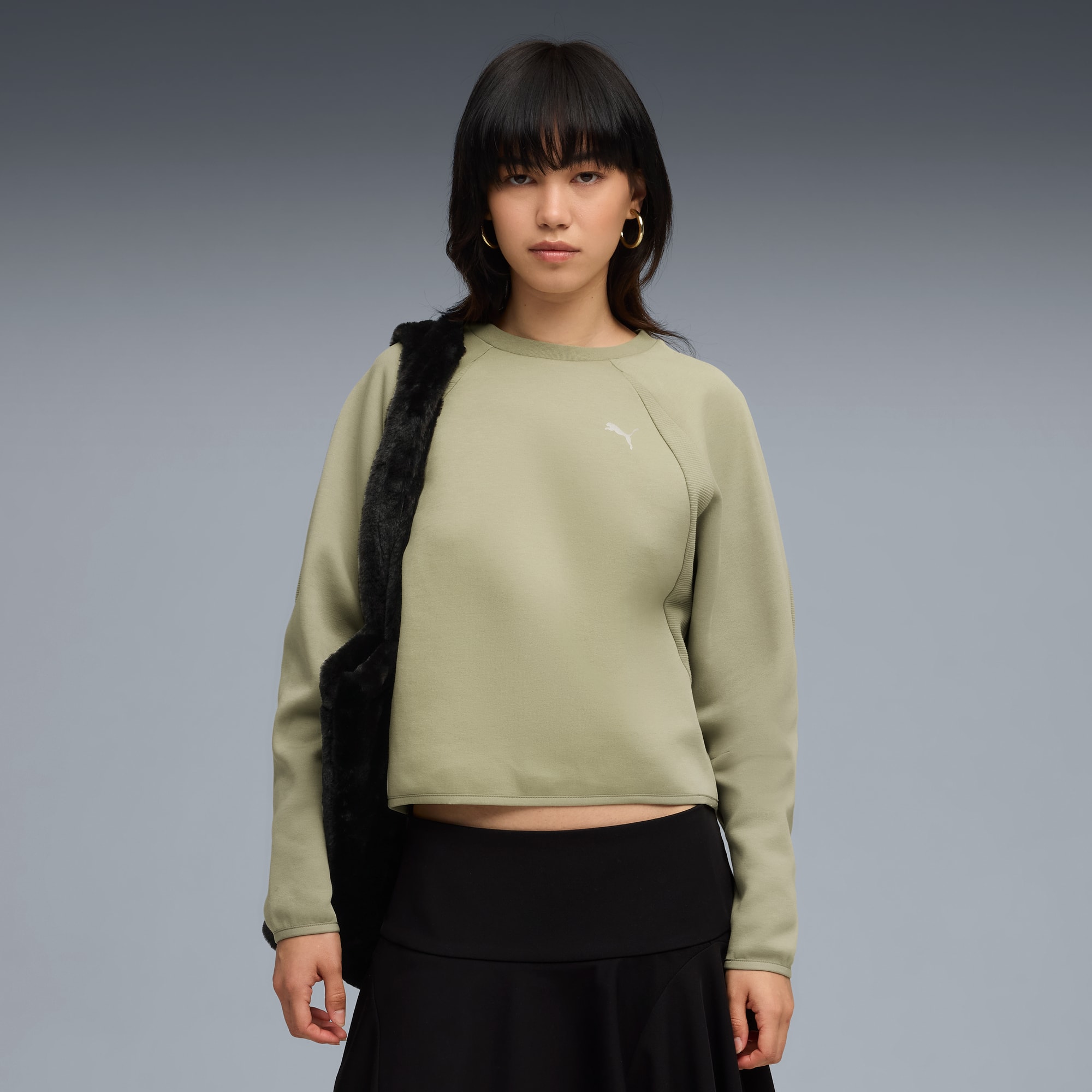 Su sfondo grigio, modella posa con felpa oliva, gonna nera e borsa soffice.