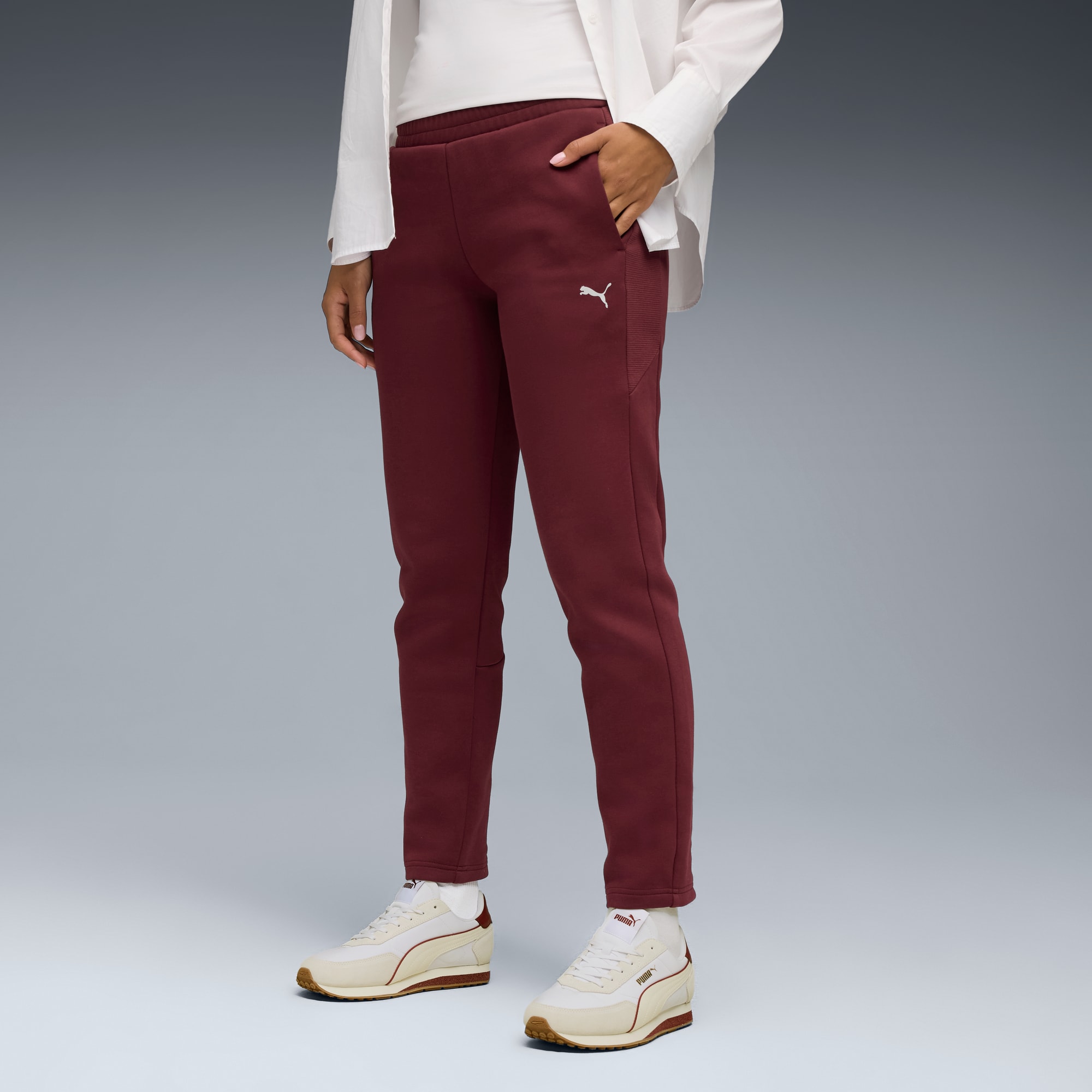 Tegen een grijze achtergrond poseert een model in bordeauxrode broek en witte sneakers.