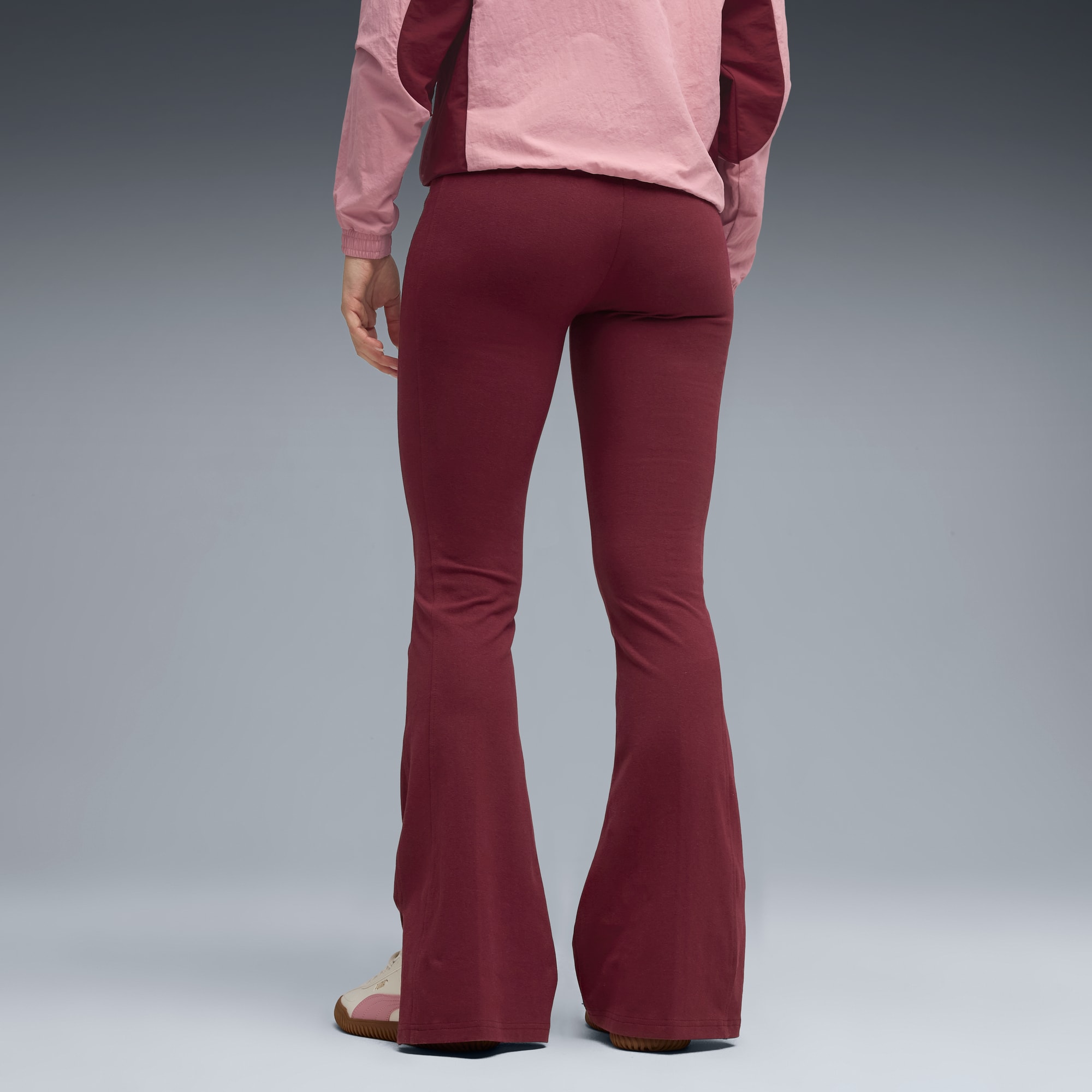 Ein Model steht vor grauem Hintergrund, trägt ein pinkes Oberteil, bordeauxrote Schlaghosen und Turnschuhe.