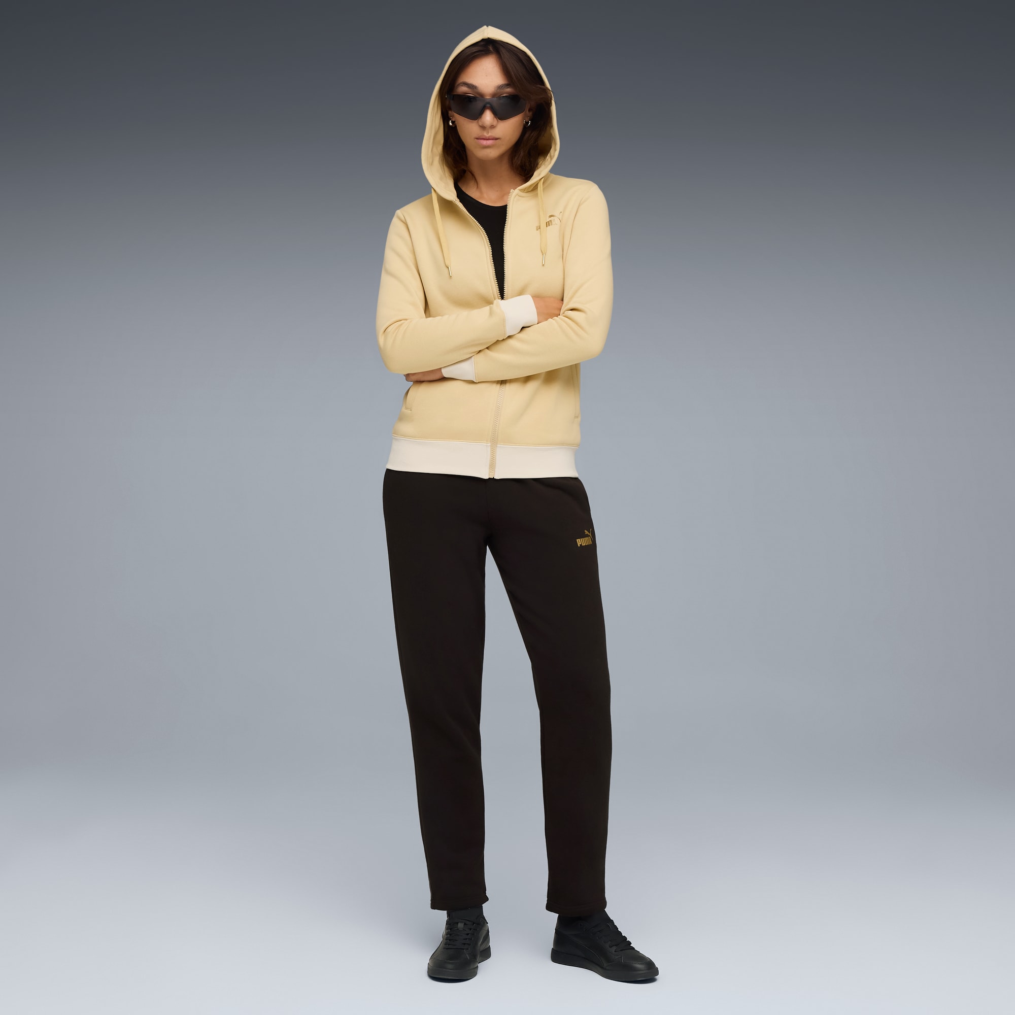 Op grijs staat een model met een jas met capuchon, een donkere broek en zonnebril.