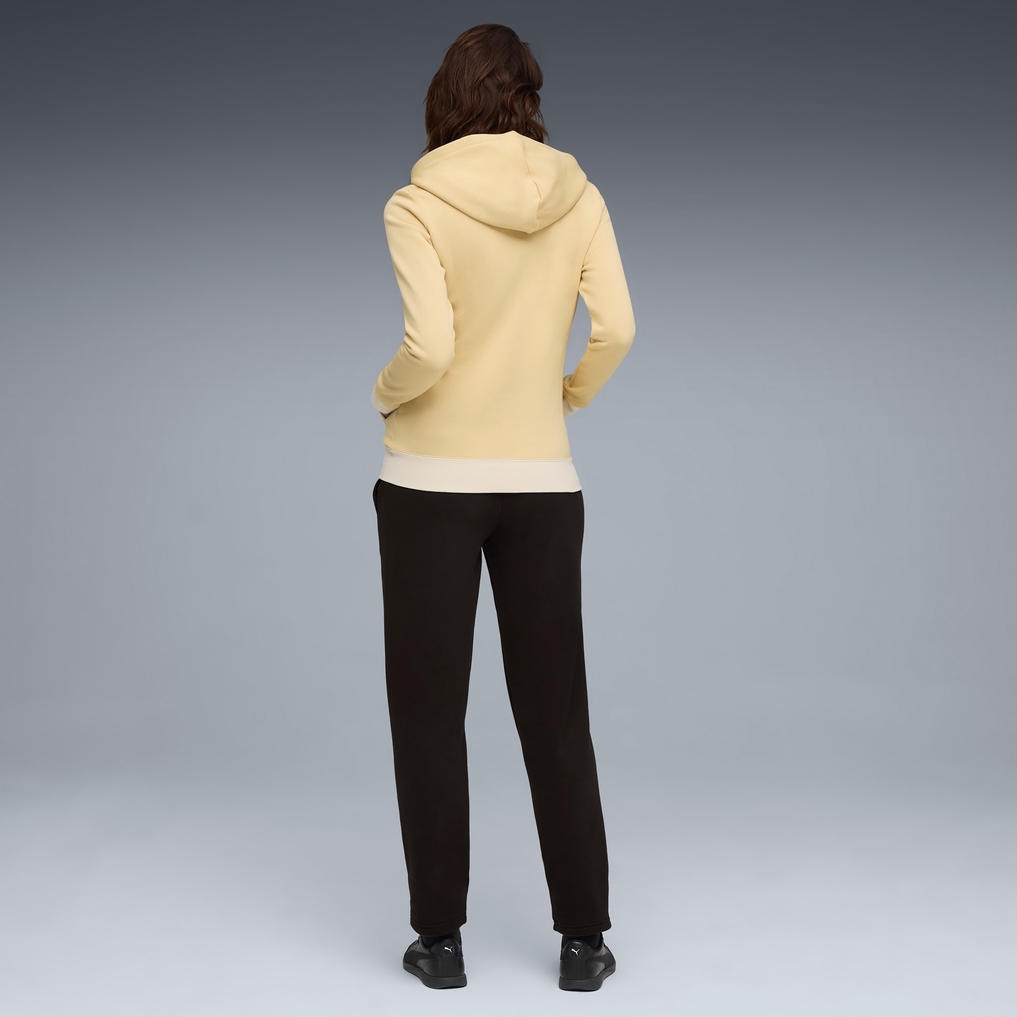 Van achteren gezien, poseert een model tegen grijs, in een gele hoodie en donkere broek.
