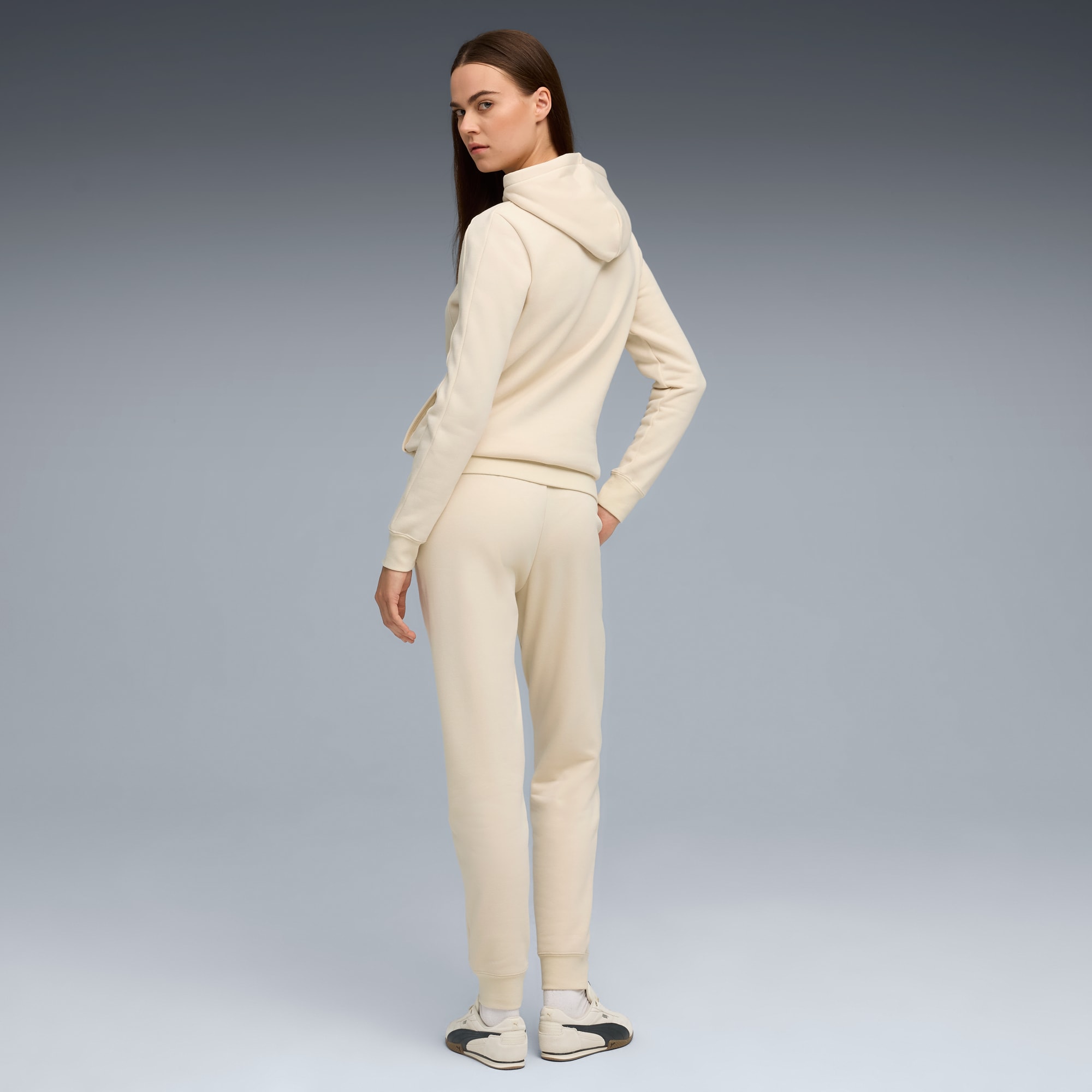 Vor grauem Hintergrund blickt ein Model zurück, trägt cremefarbenen Hoodie, Jogginghose und Turnschuhe.