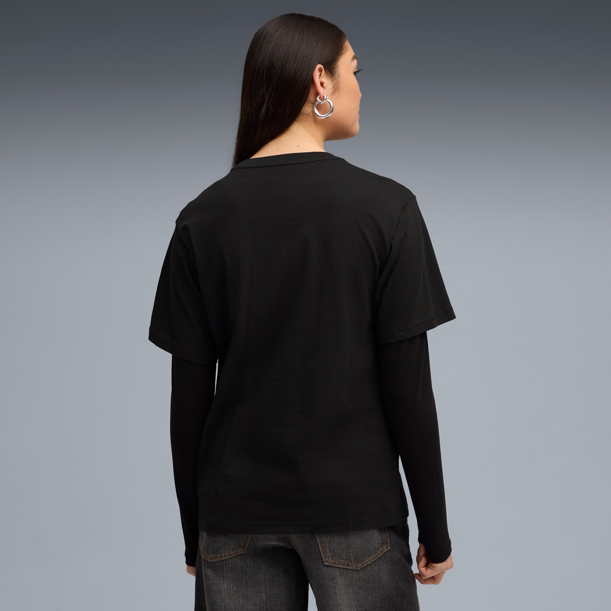 Vor grauem Hintergrund trägt ein Model ein schwarzes T-Shirt, Langarmshirt und dunkle Jeans.