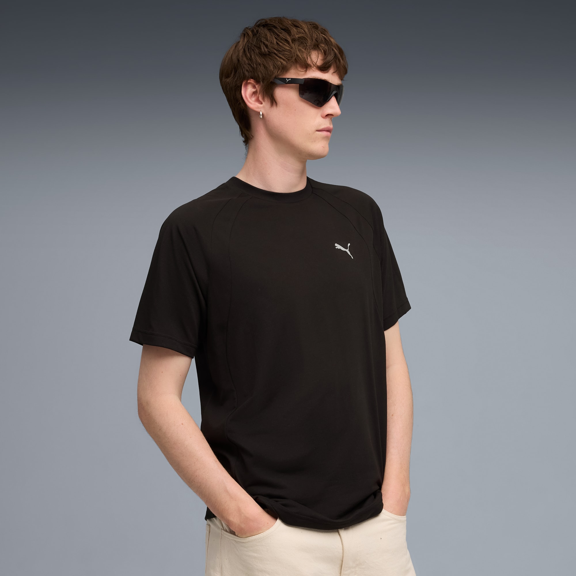 Ein Model posiert vor grauem Hintergrund mit schwarzem T-Shirt, Sonnenbrille und heller Hose.