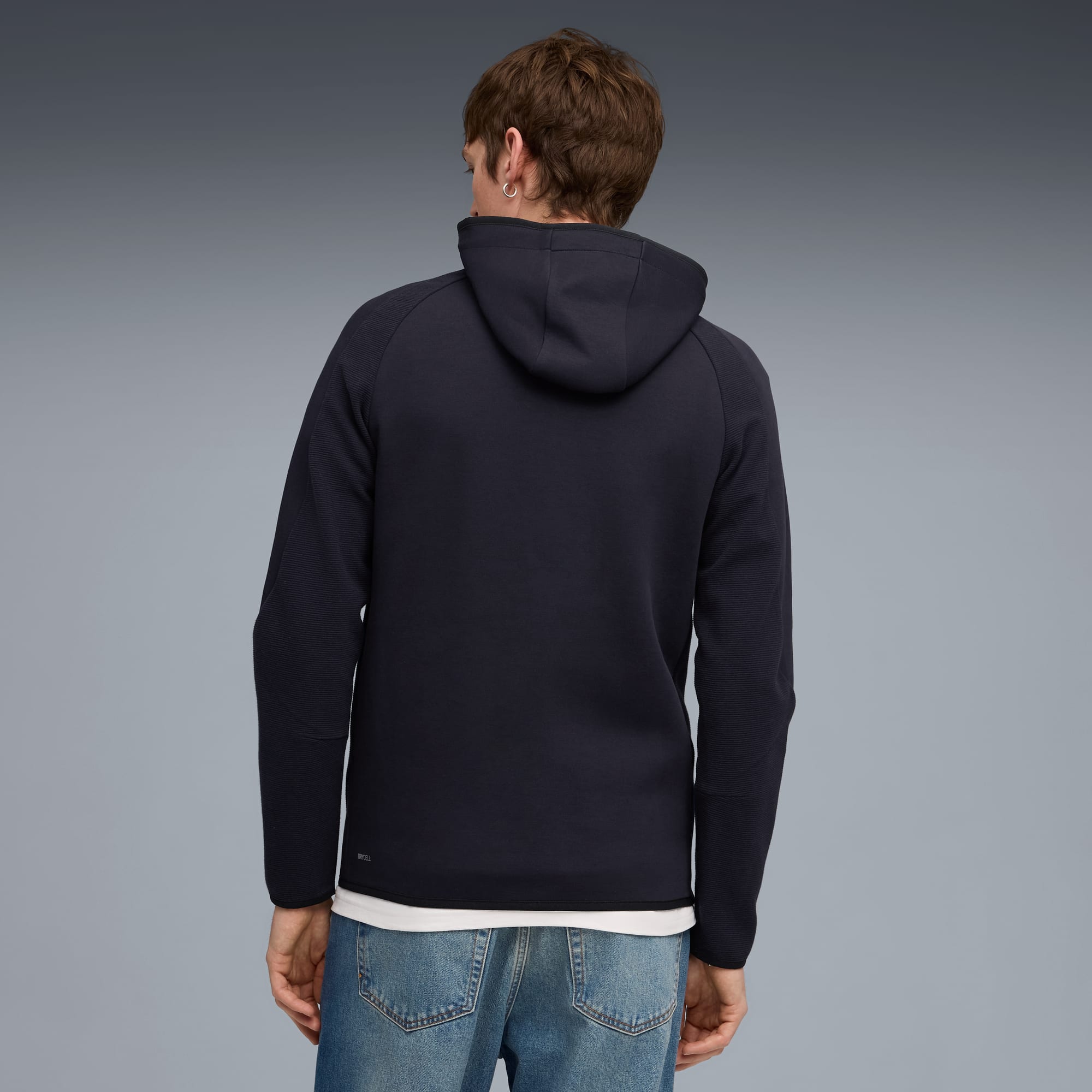 Een persoon staat met de rug naar de camera, draagt een donkere hoodie en jeans, tegen grijs.