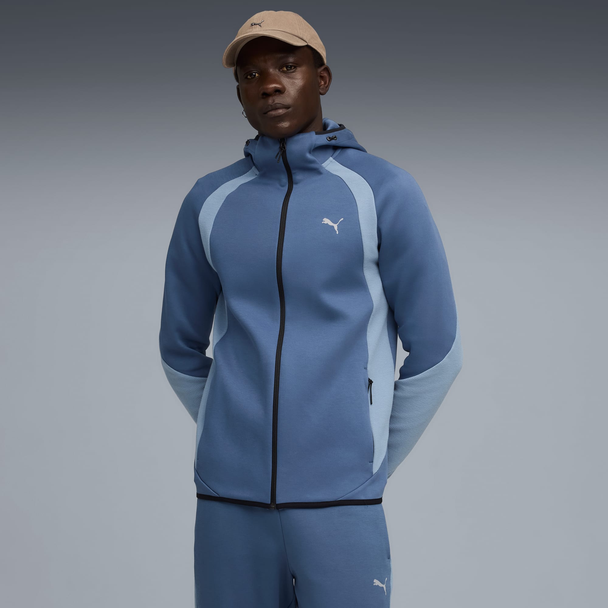 Ein Model steht vor grauem Hintergrund und trägt eine blaue Kapuzenjacke, Hose und beigefarbene Kappe.