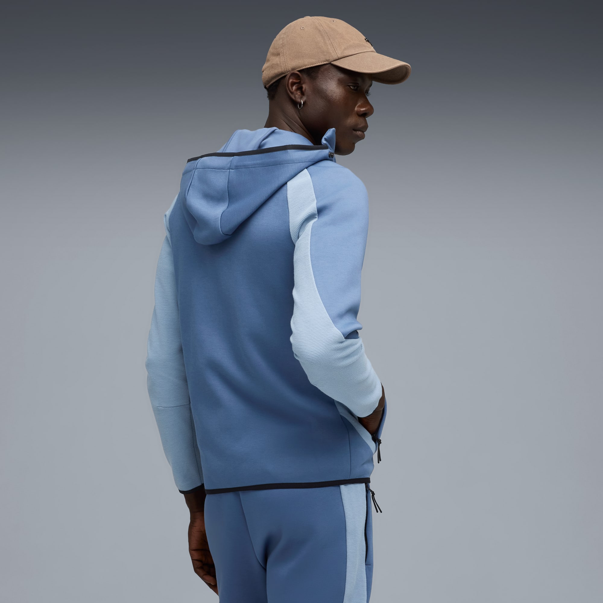 Vor grauem Hintergrund blickt ein Model nach links, mit Kappe und blauem Trainingsanzug mit Kapuze.