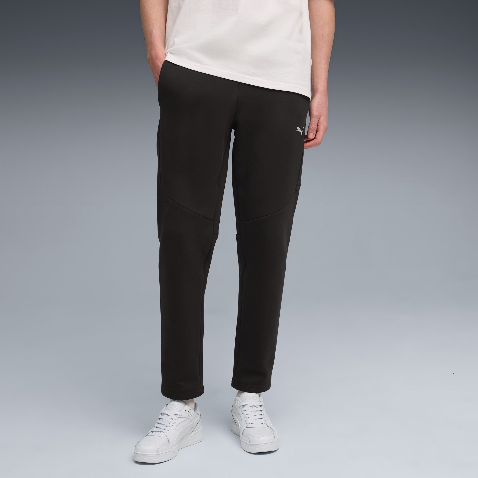 Un mannequin pose sur fond gris, portant un t-shirt blanc, un pantalon noir et des baskets blanches.