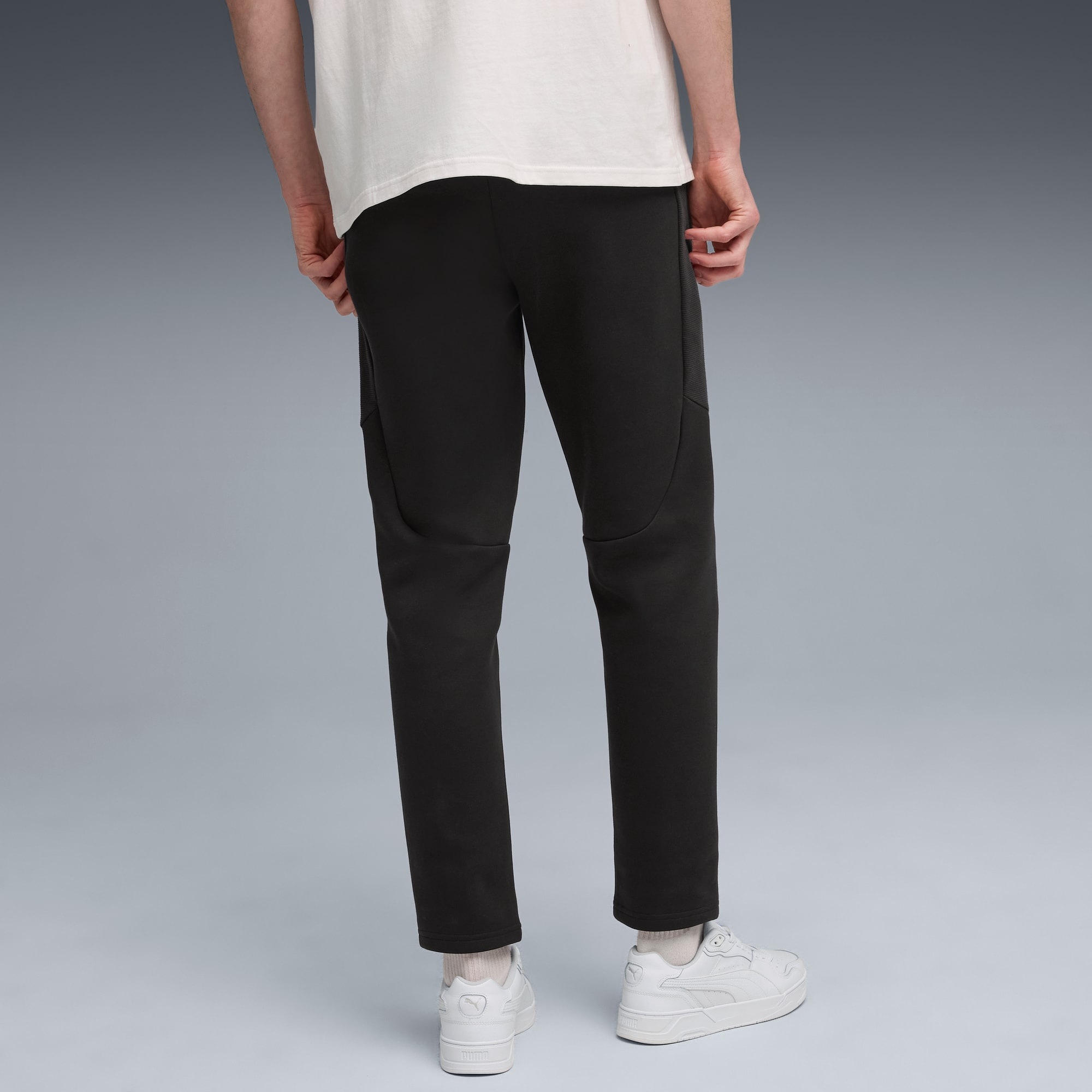Ein Model vor grauem Hintergrund trägt schwarze Hose, weißes T-Shirt und weiße Turnschuhe.