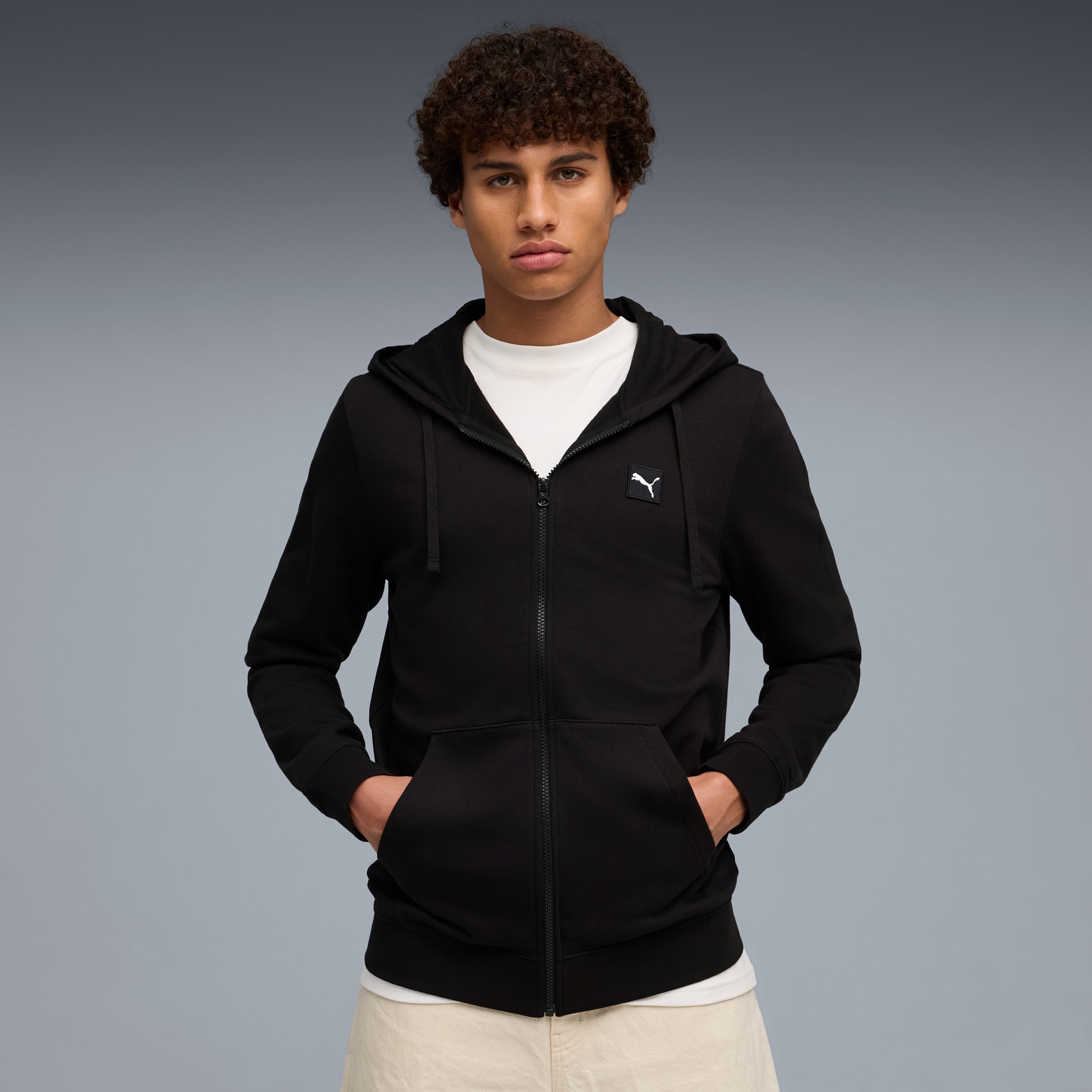 Ein Model mit Händen in den Taschen trägt einen schwarzen Reißverschluss-Hoodie über einem weißen T-Shirt.