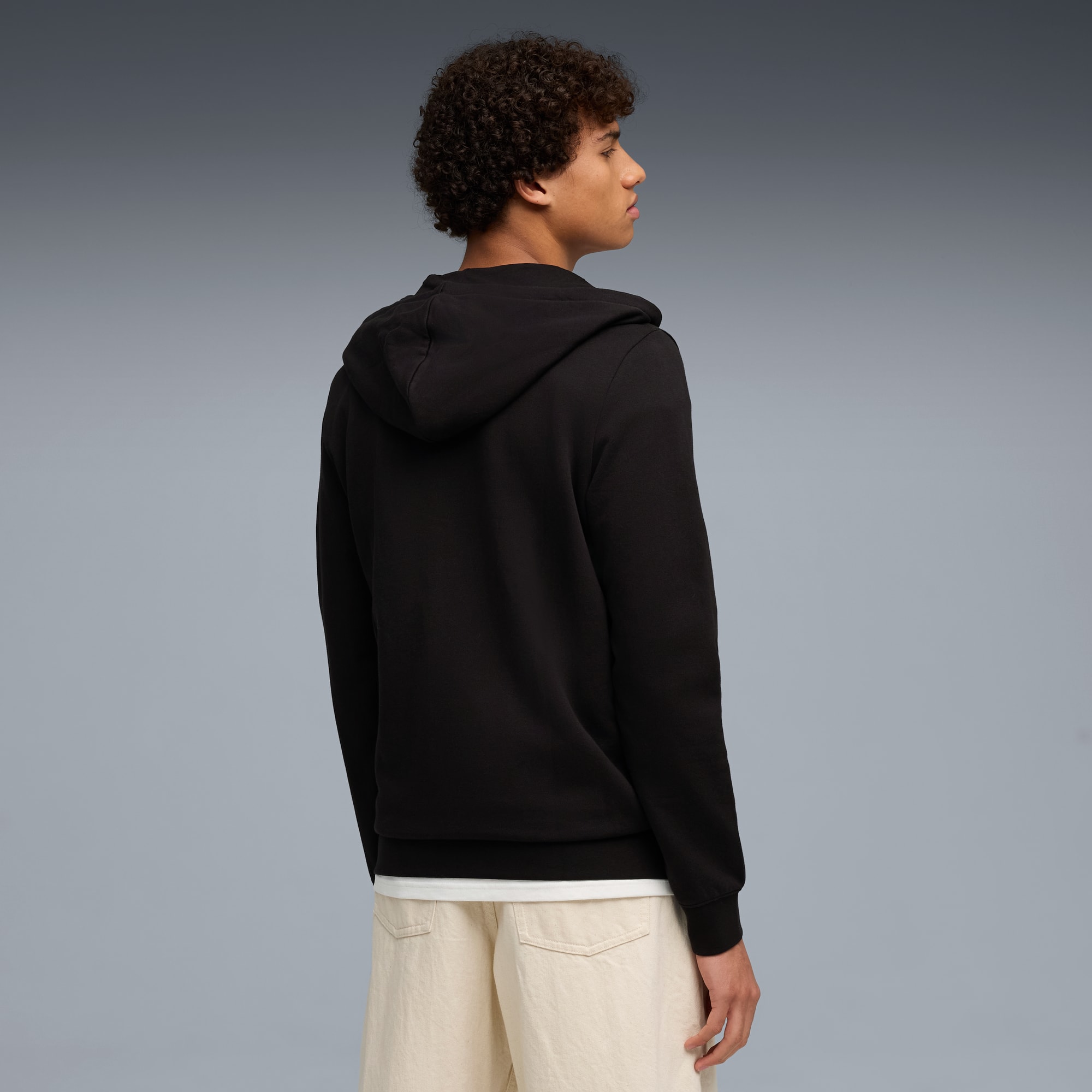 Vor grauem Hintergrund trägt ein Model eine schwarze Kapuzenjacke und beigefarbene Shorts.
