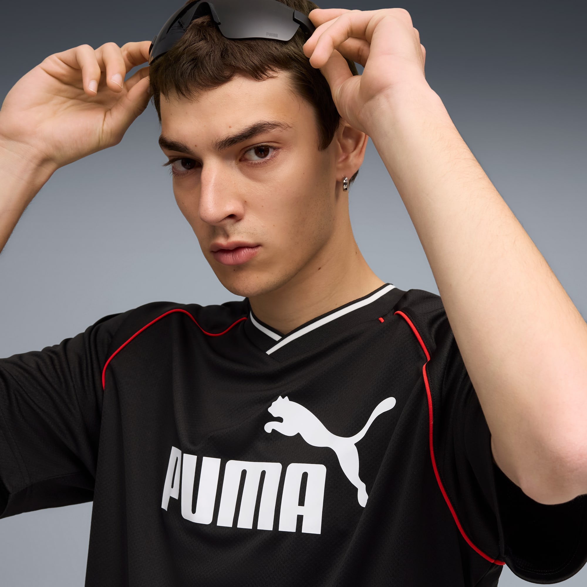 Ein Model justiert eine Sonnenbrille und tr&auml;gt ein schwarzes Puma-T-Shirt vor grauem Hintergrund.