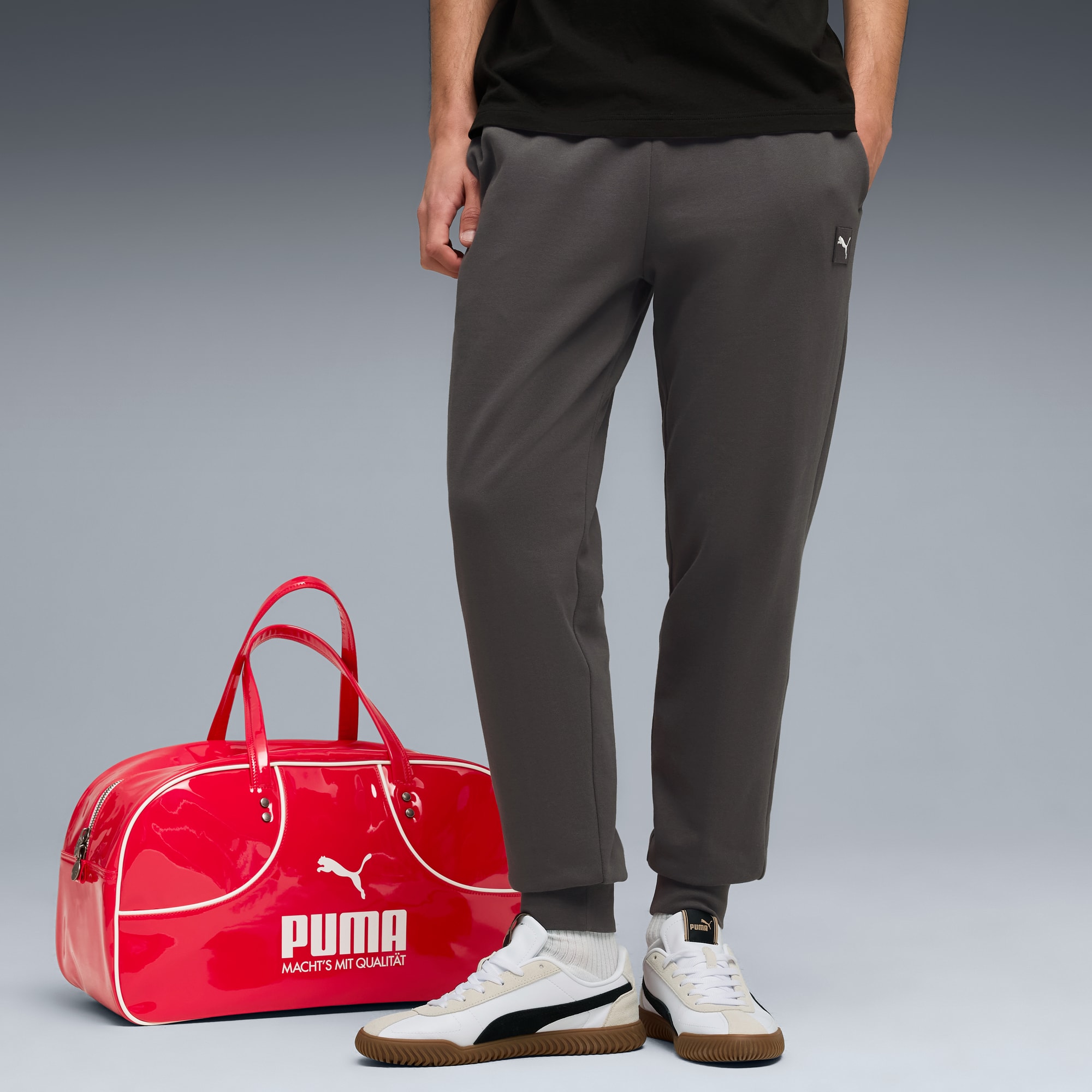 Op grijs draagt een model een grijze broek en sneakers naast een glimmende rode weekendtas.