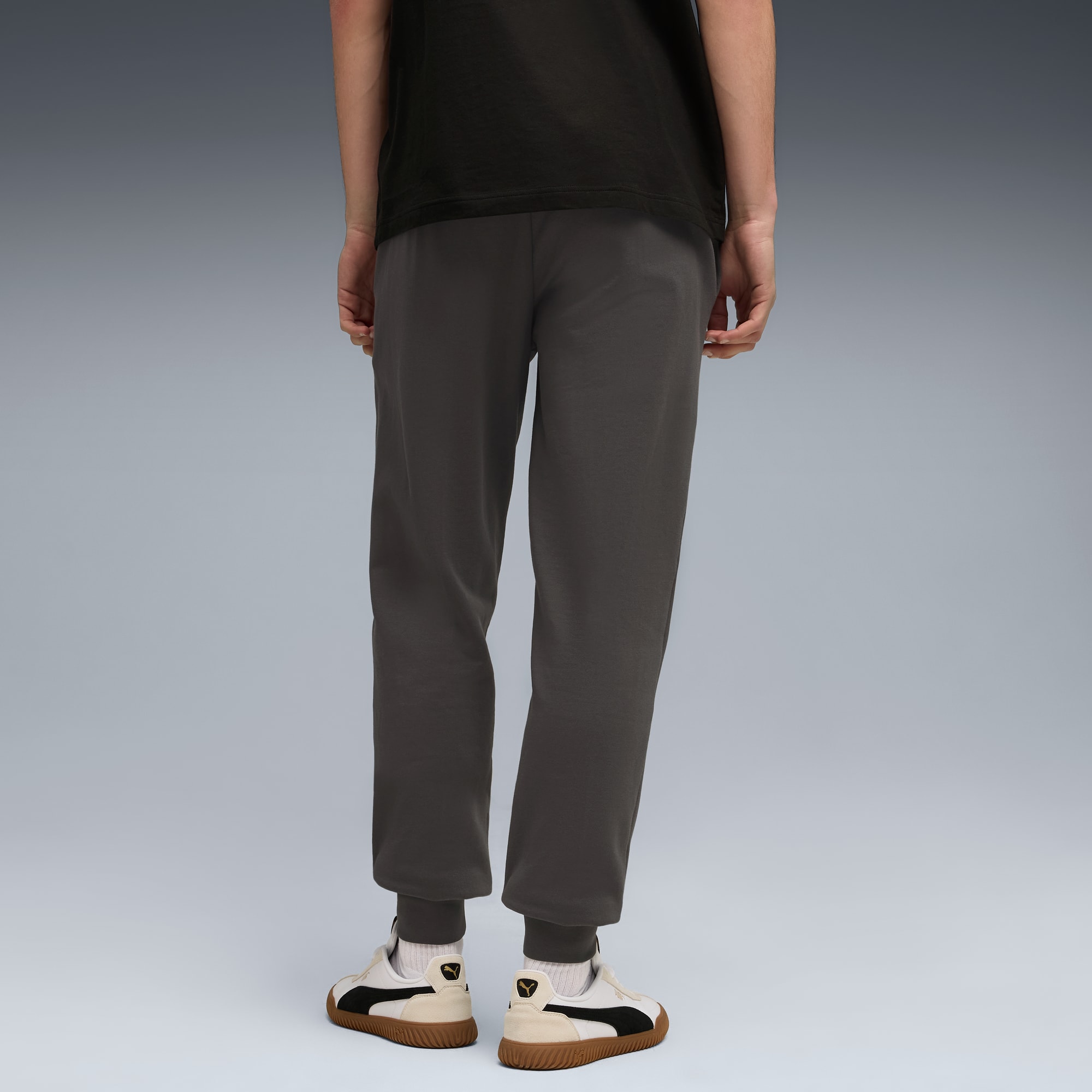 Een model staat met de rug naar de camera op grijs, gekleed in een donker t-shirt, grijze broek en crème-zwarte sneakers.