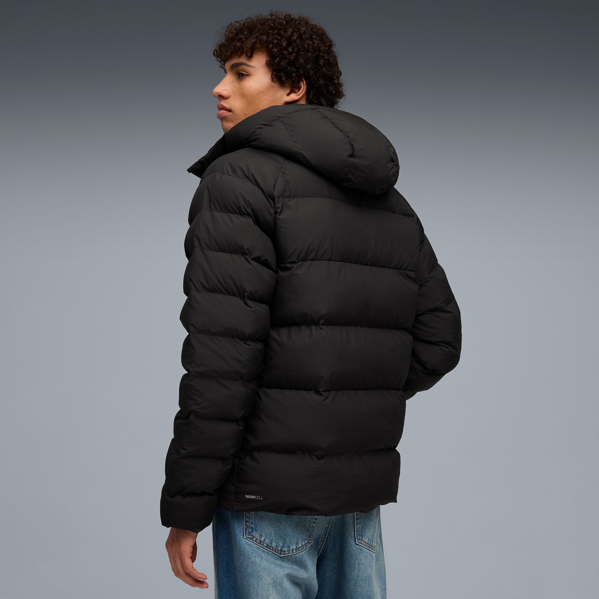 Ein Model posiert vor grauem Hintergrund mit schwarzer Kapuzen-Pufferjacke und Jeans.