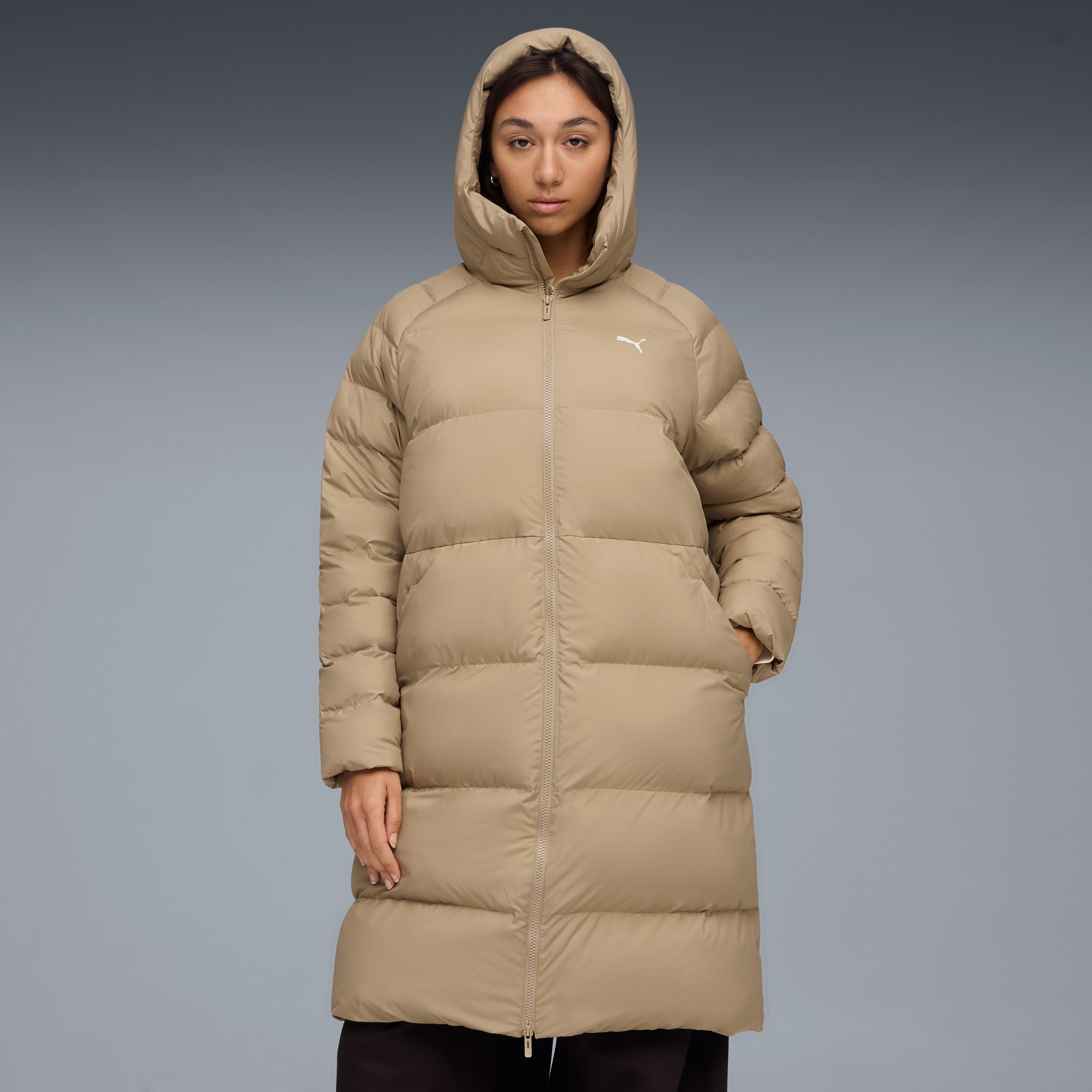 Su sfondo grigio, modella in lungo piumino beige con cappuccio e logo bianco.