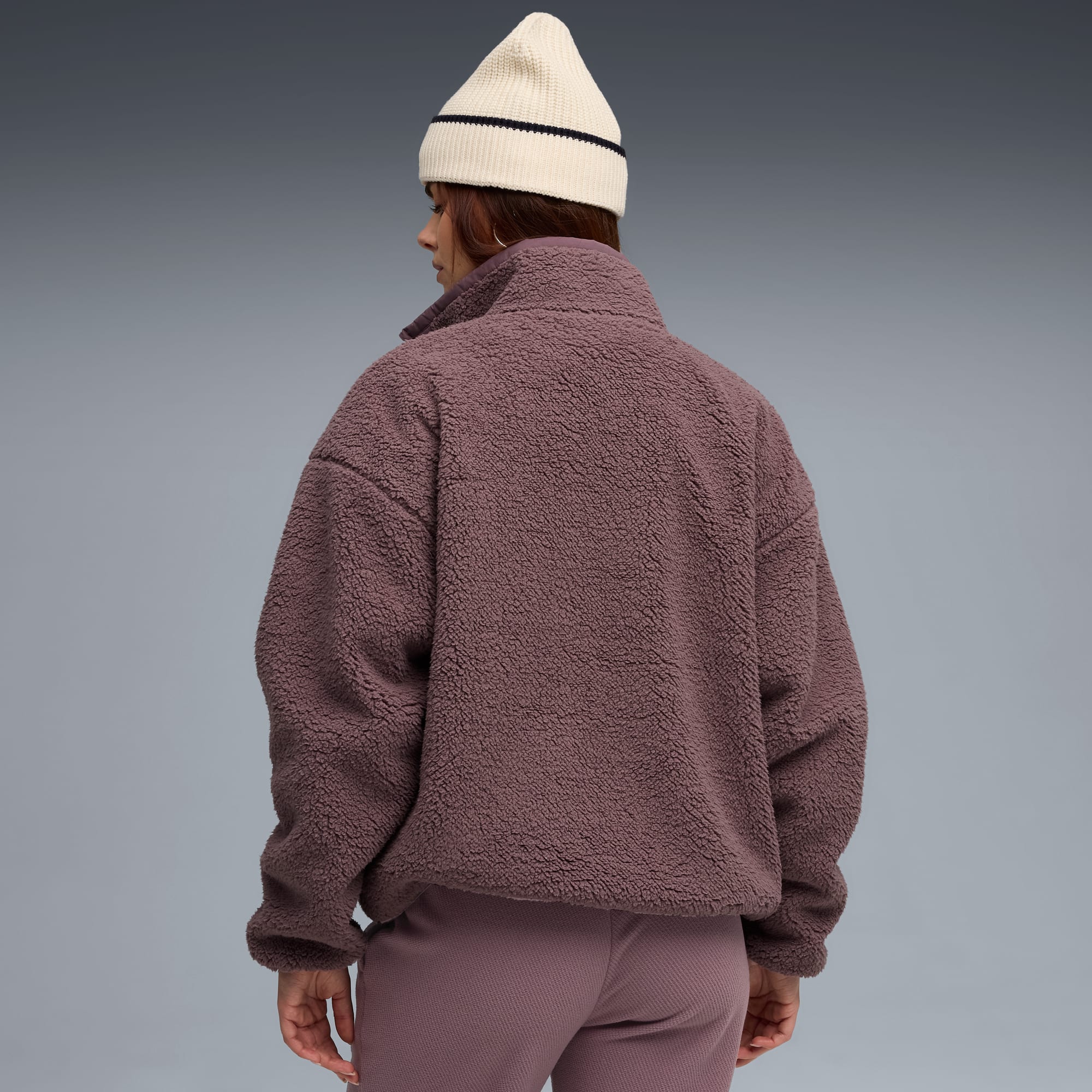 Una modella di spalle su sfondo grigio indossa una giacca in pile color malva, pantaloni abbinati e un berretto a righe.