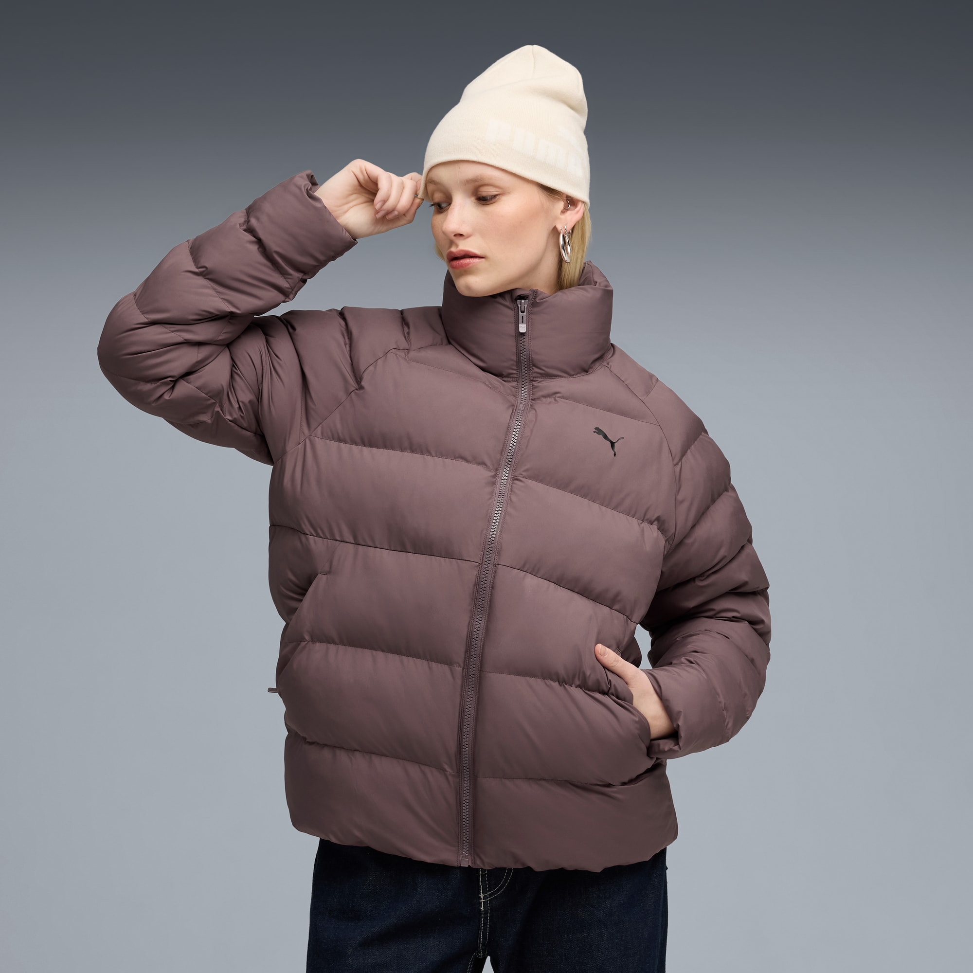 Modella posa su sfondo grigio con piumino marrone e berretto crema.