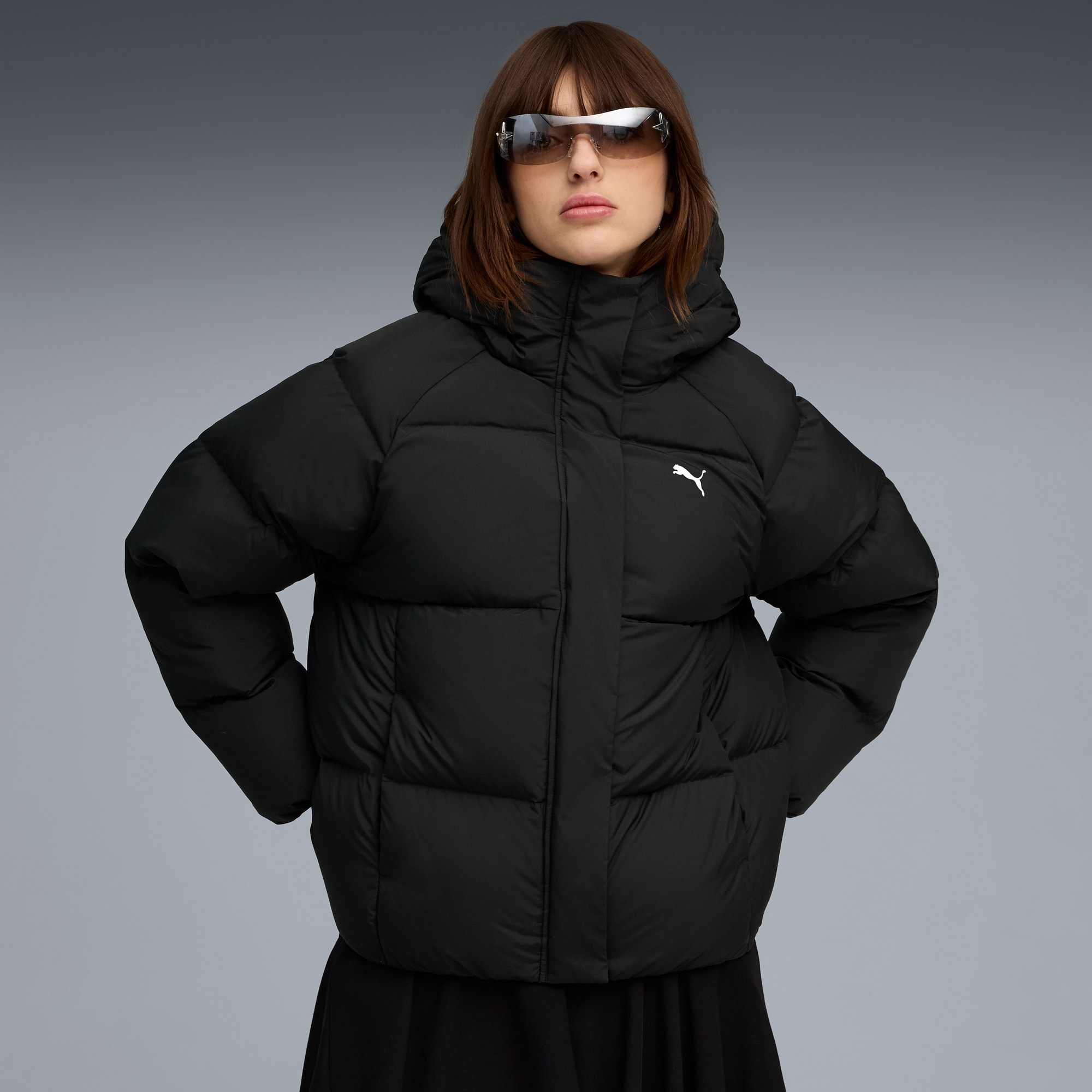 Sobre un fondo gris, una modelo posa con una chaqueta acolchada negra y gafas de sol.