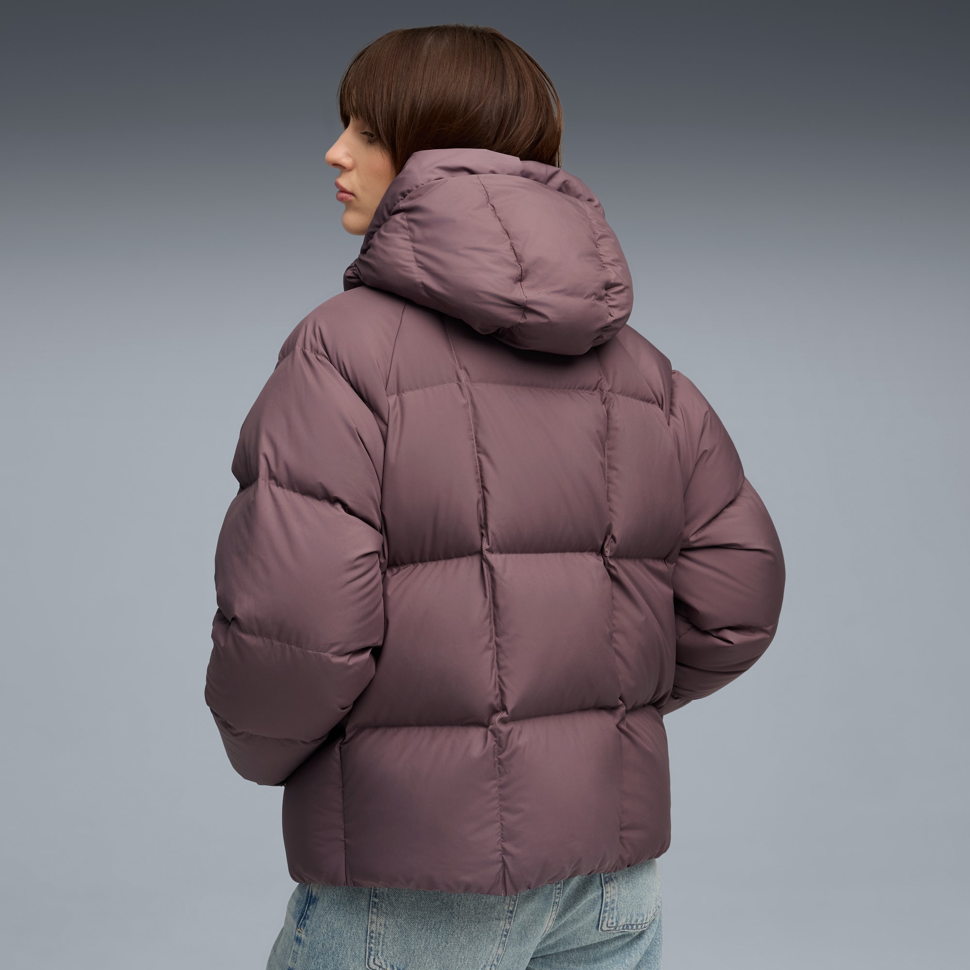 Ein Model posiert vor grauem Hintergrund mit einem mauvefarbenen Puffer-Mantel und Jeans.