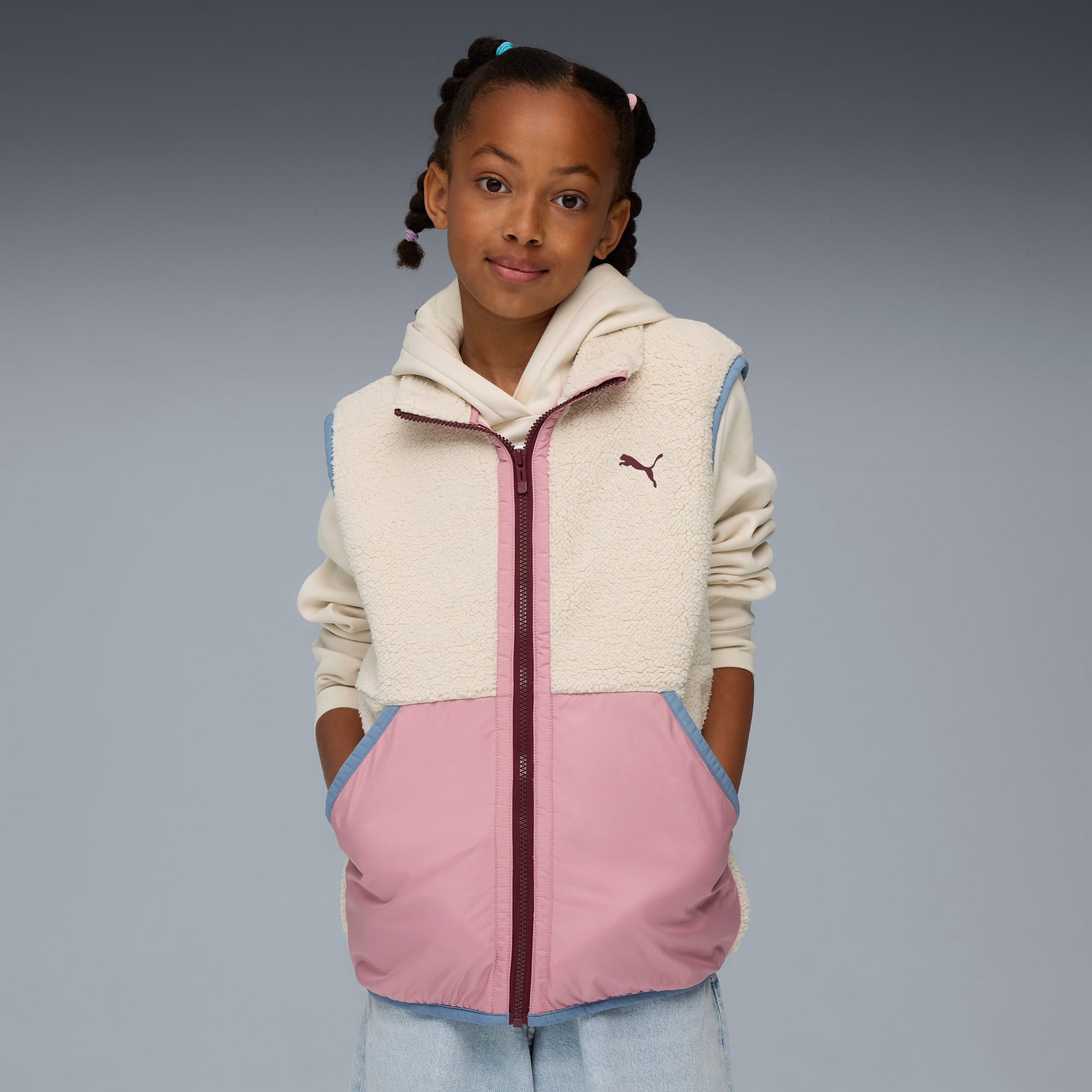 Un modello bambino posa su uno sfondo grigio, indossando una felpa con cappuccio e un gilet color crema e rosa.