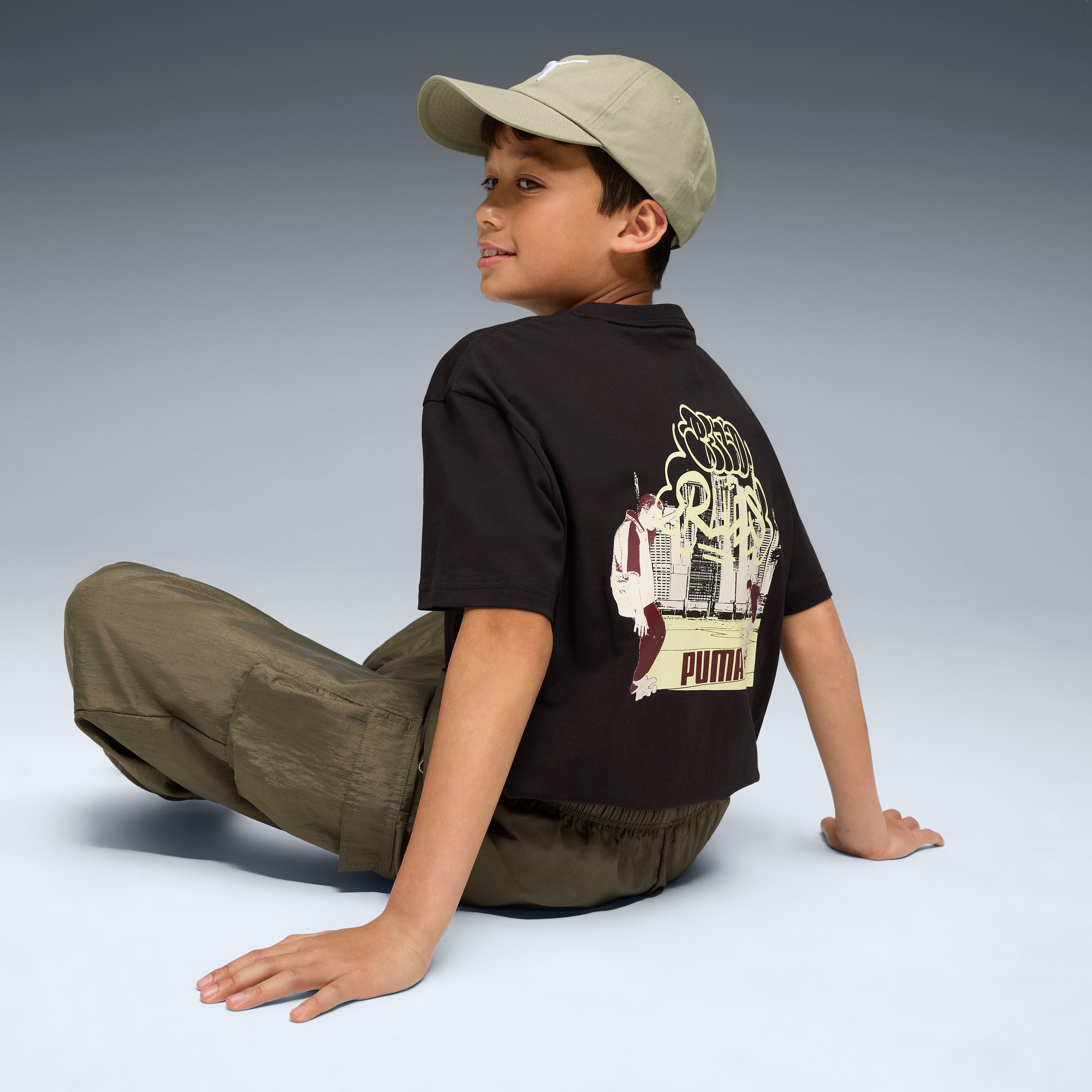 Een kind zit tegen een grijze achtergrond, met een pet, bedrukt T-shirt en broek.