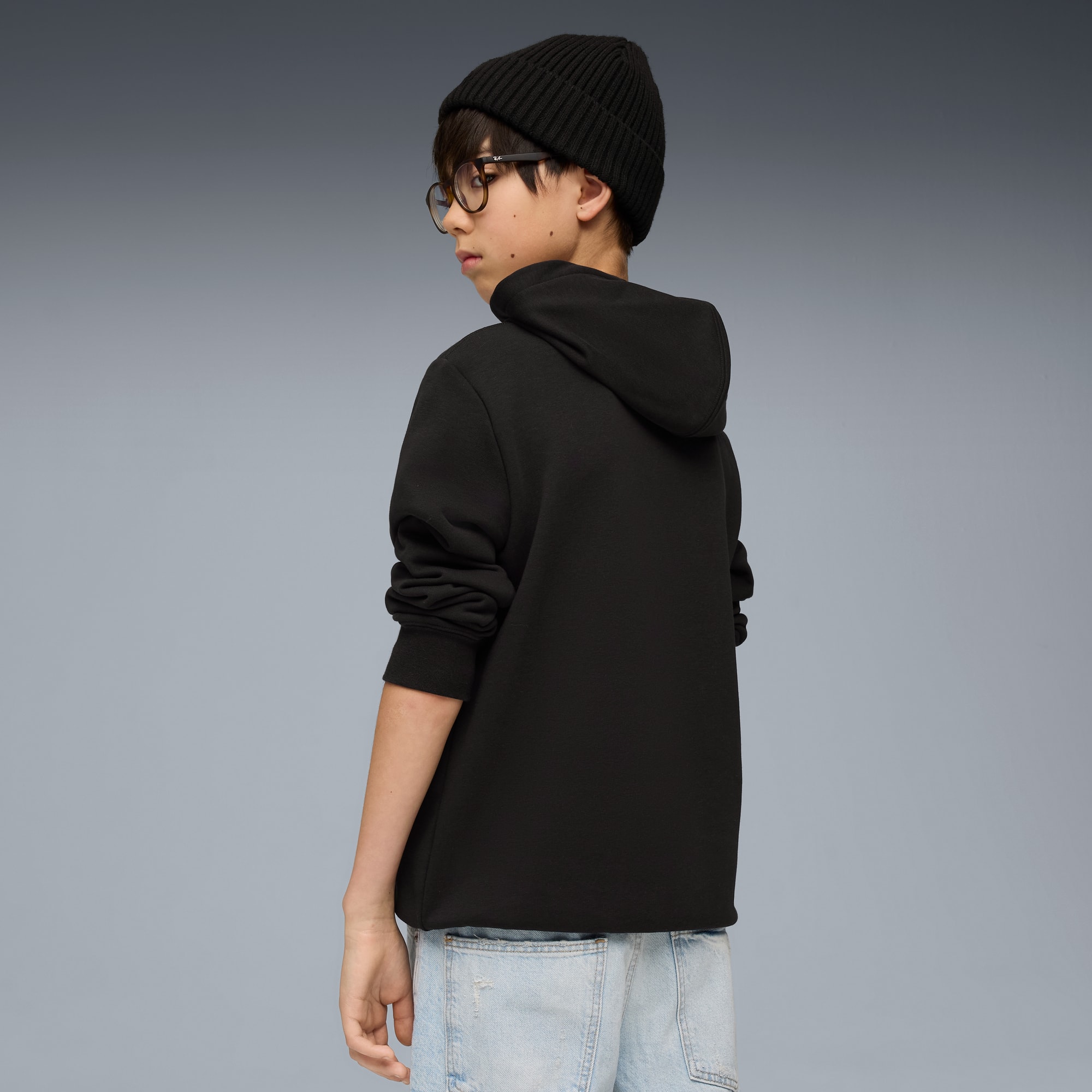 Tegen een grijze achtergrond kijkt een model achterom, met een zwarte hoodie, muts en bril.