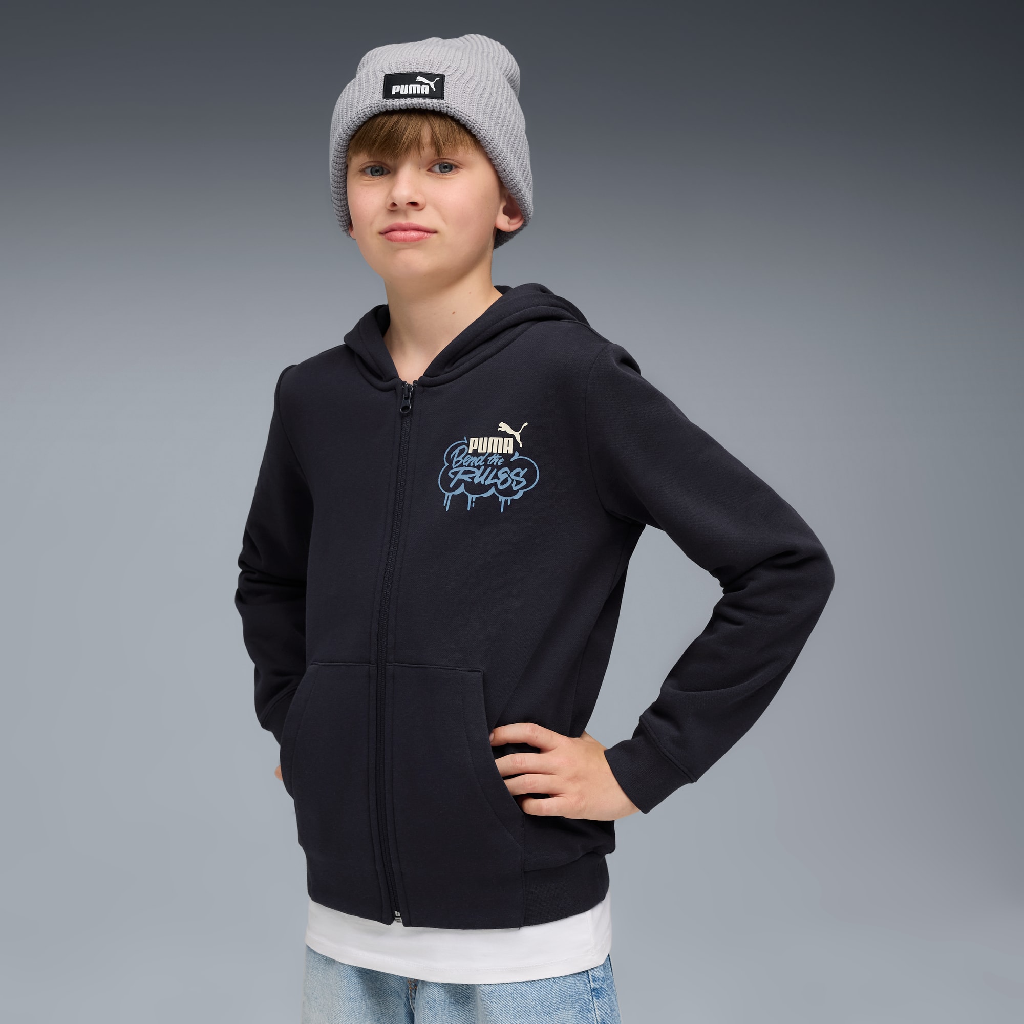 Op grijs poseert een jongen met een grijze beanie en een zwart vest met rits en print.