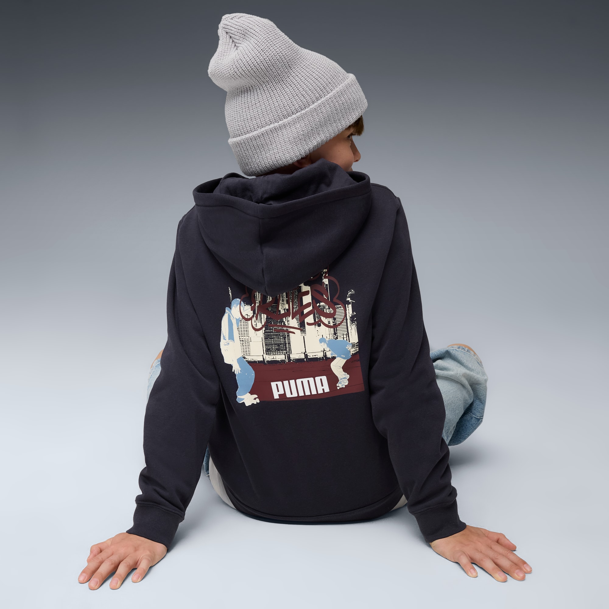 Een model op een grijze achtergrond draagt een muts en een bedrukte Puma-hoodie.