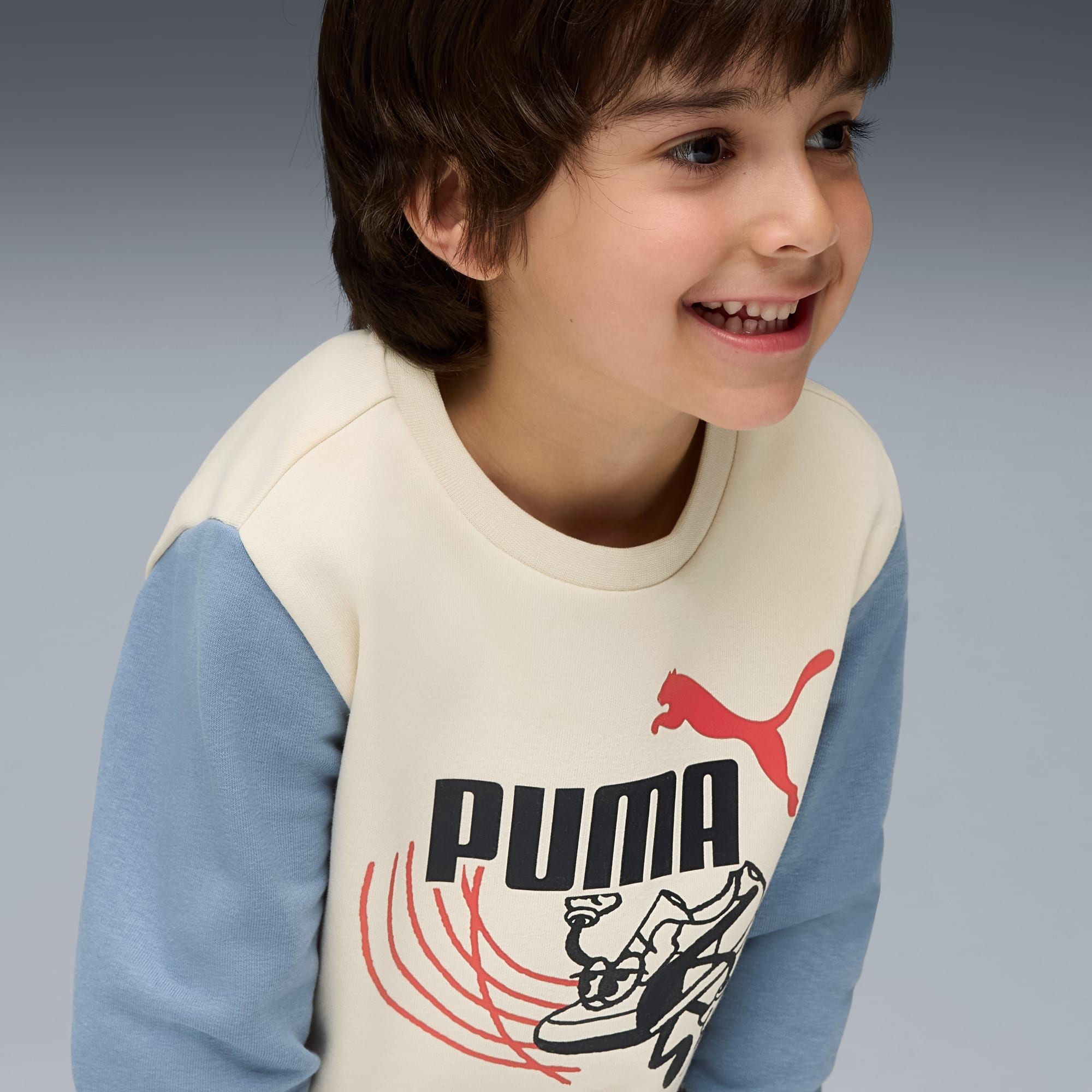 Ein lächelndes Kind mit Puma-Sweatshirt vor grauem Hintergrund.
