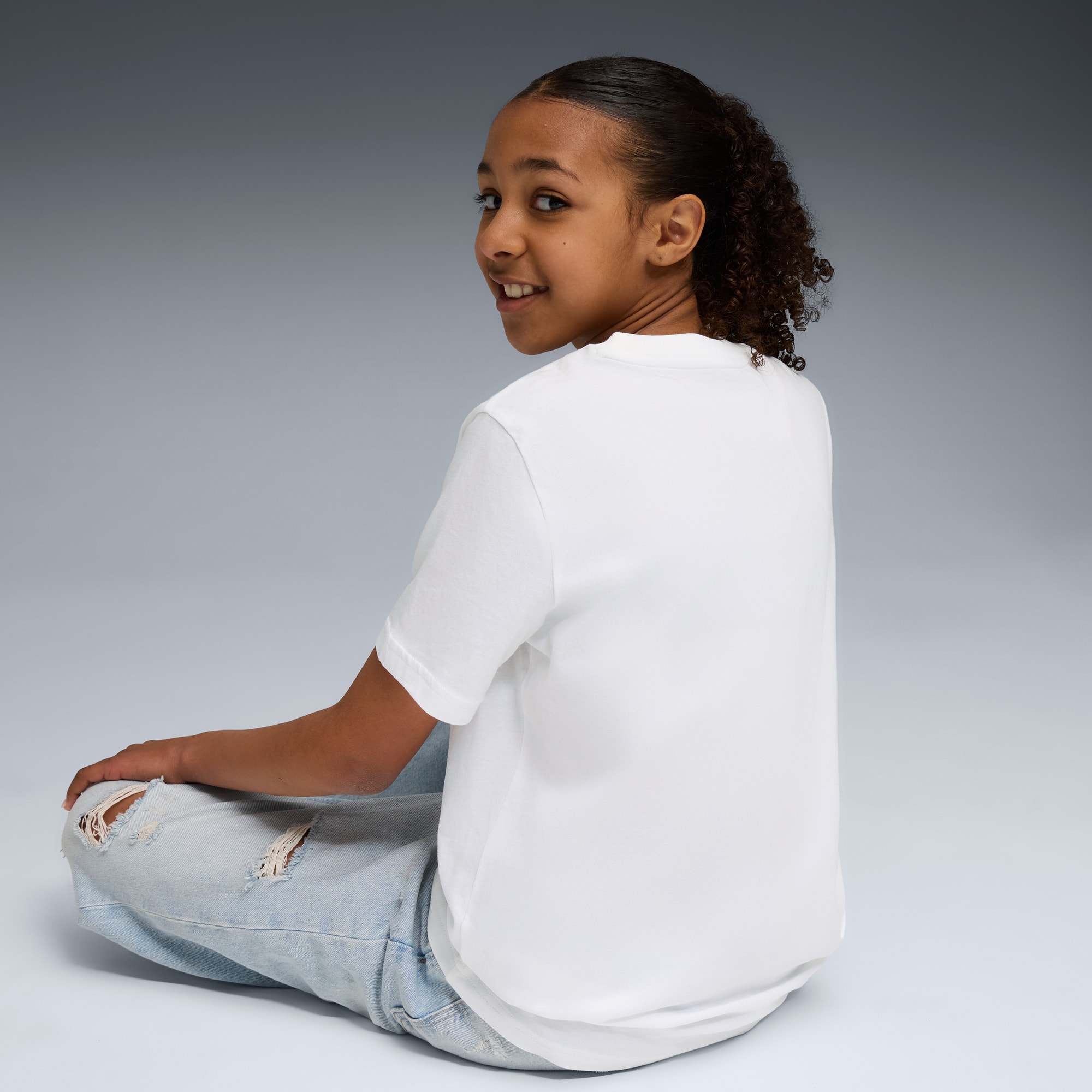 Op grijs zit een model gekruist met een wit T-shirt en gescheurde lichtblauwe jeans.
